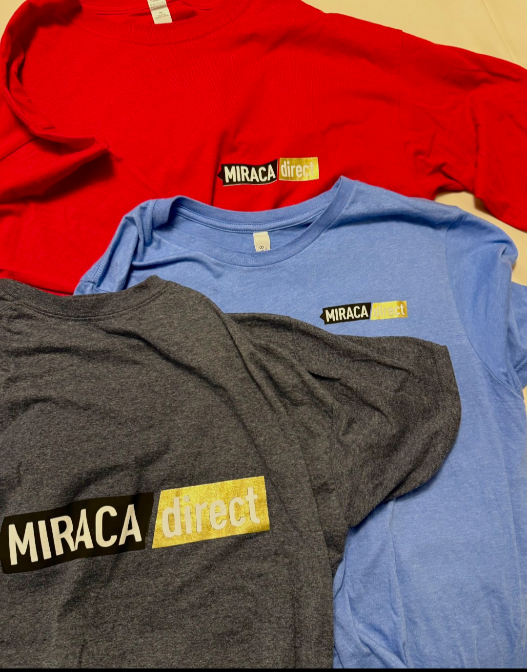 Three t-shirts in red, blue, and dark gray colors, each with a "MIRACA direct" logo on the front, laid out on a flat surface.