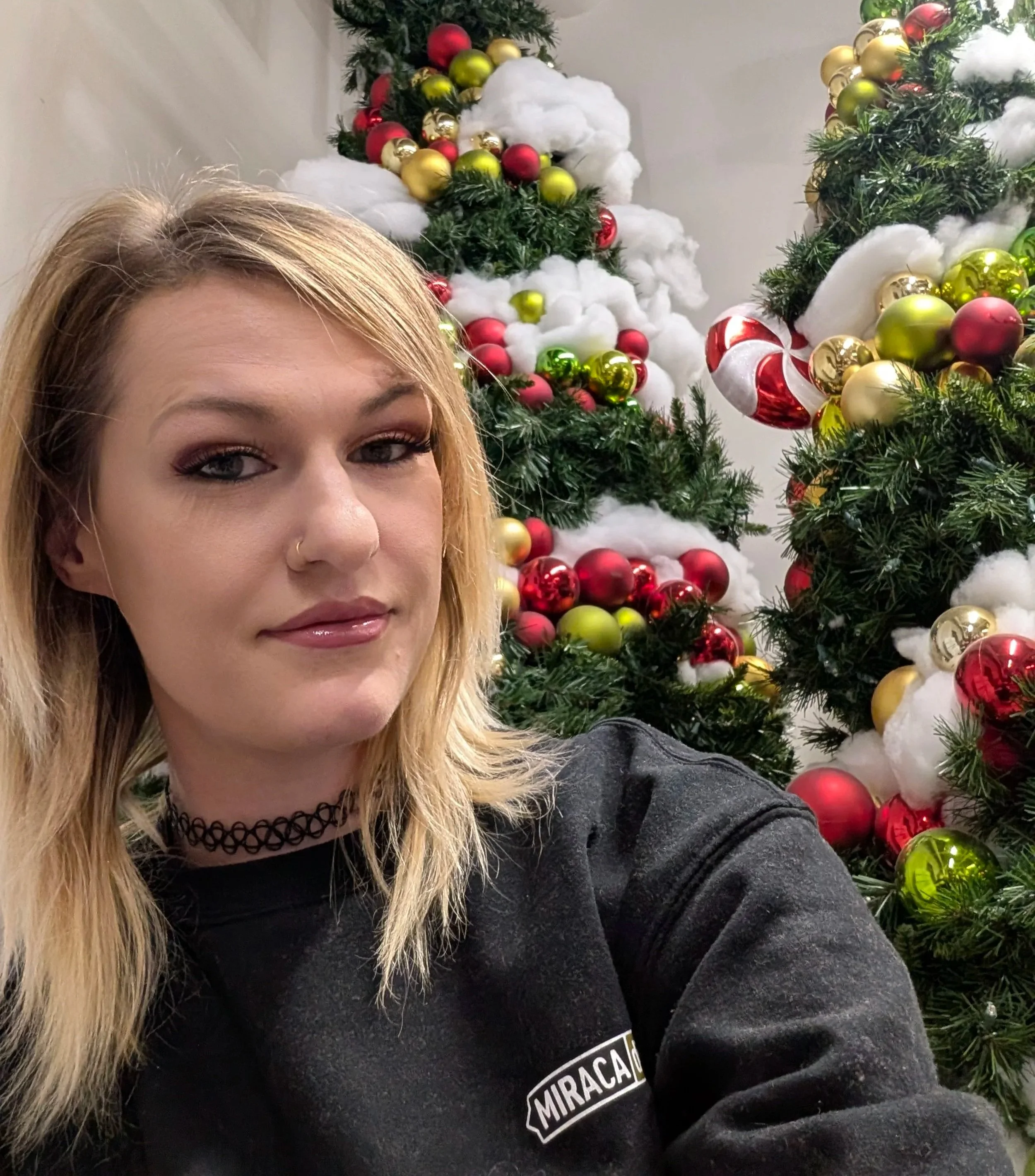 A woman taking a selfie in front of decorated Christmas trees with red, gold, and green ornaments, and cotton-like snow decorations.