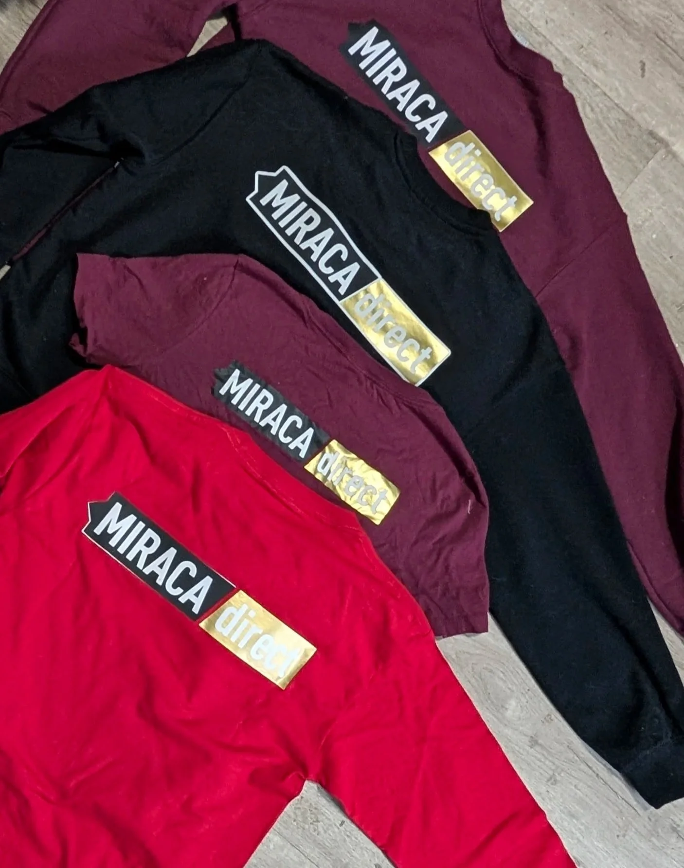 Three hoodies in red, black, and maroon colors with 'MIRACA direct' logo on the front, laid out on a wooden floor.