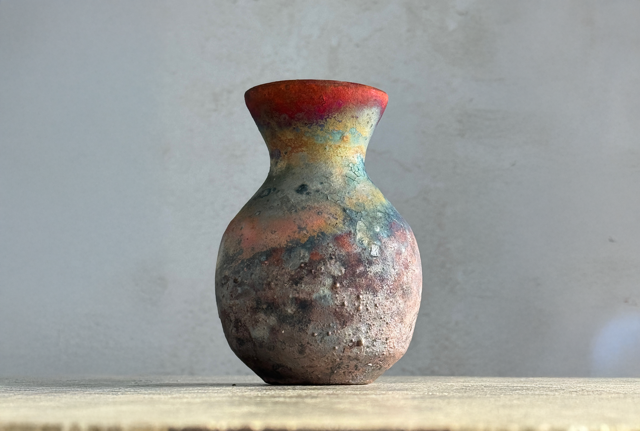 A rustic, weathered ceramic vase with a narrow neck and bulbous body, featuring a multicolored, textured surface with shades of red, blue, yellow, and black, set on a wooden surface against a plain, light gray wall.
