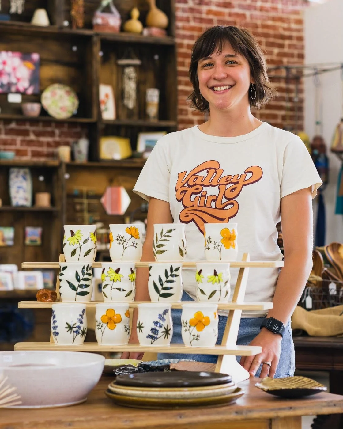 A smiling woman wearing a white t-shirt with 'Valley Girl' written on it, standing behind a display of ceramic cups with floral designs on a wooden rack inside a store.