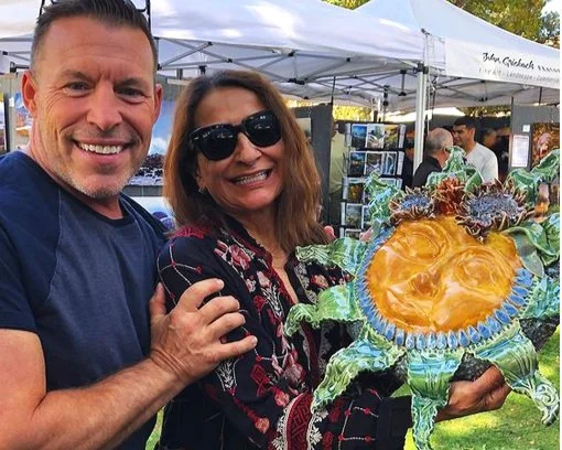 A smiling man and woman holding a colorful crab-shaped glass art piece at an outdoor event with booths and tents in the background.