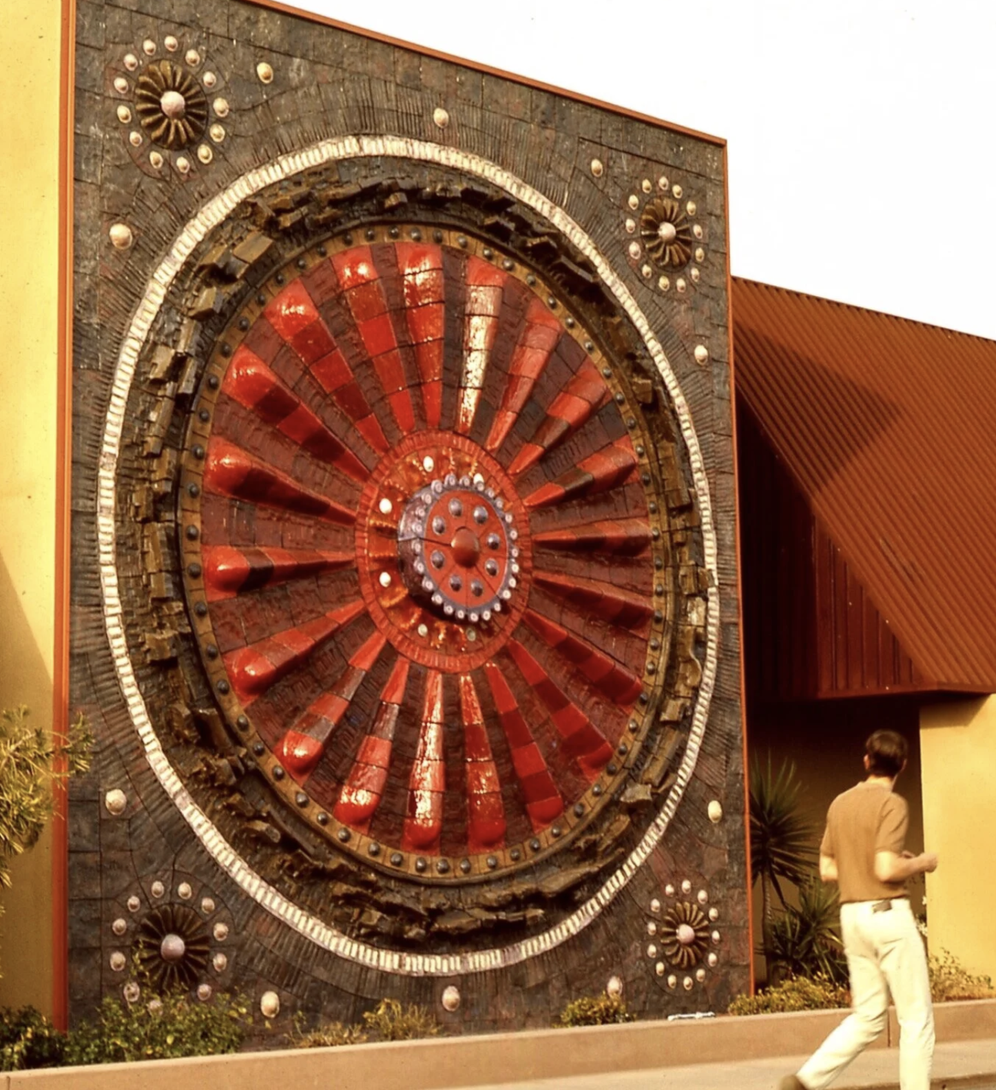 A large, decorative, circular wall sculpture with a radiating pattern of red and brown segments, adorned with small circular elements, set against a brick wall exterior of a building. A person is walking nearby.