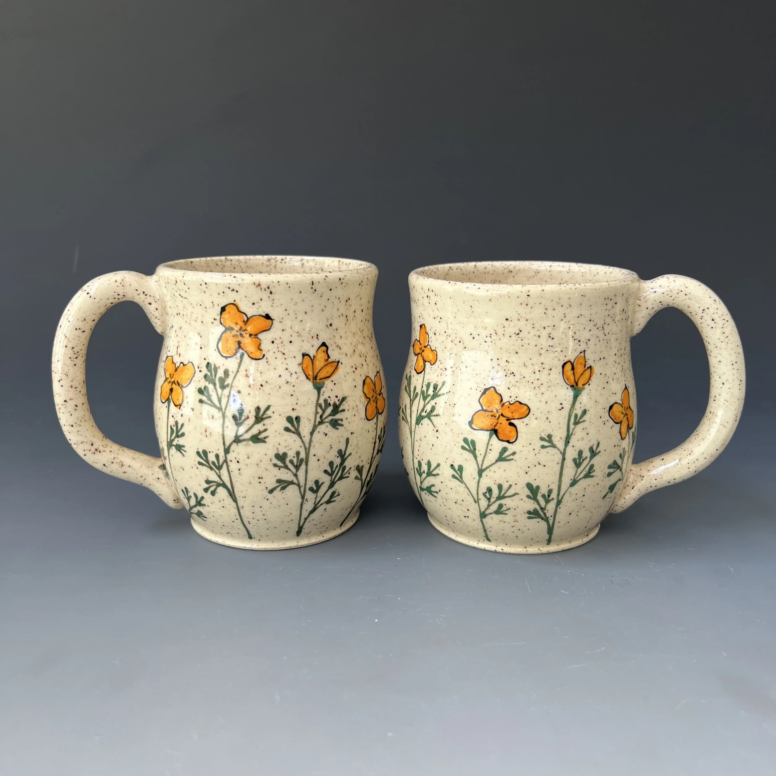 Two ceramic mugs with orange flowers and green leaves painted on them, placed against a plain gray background.