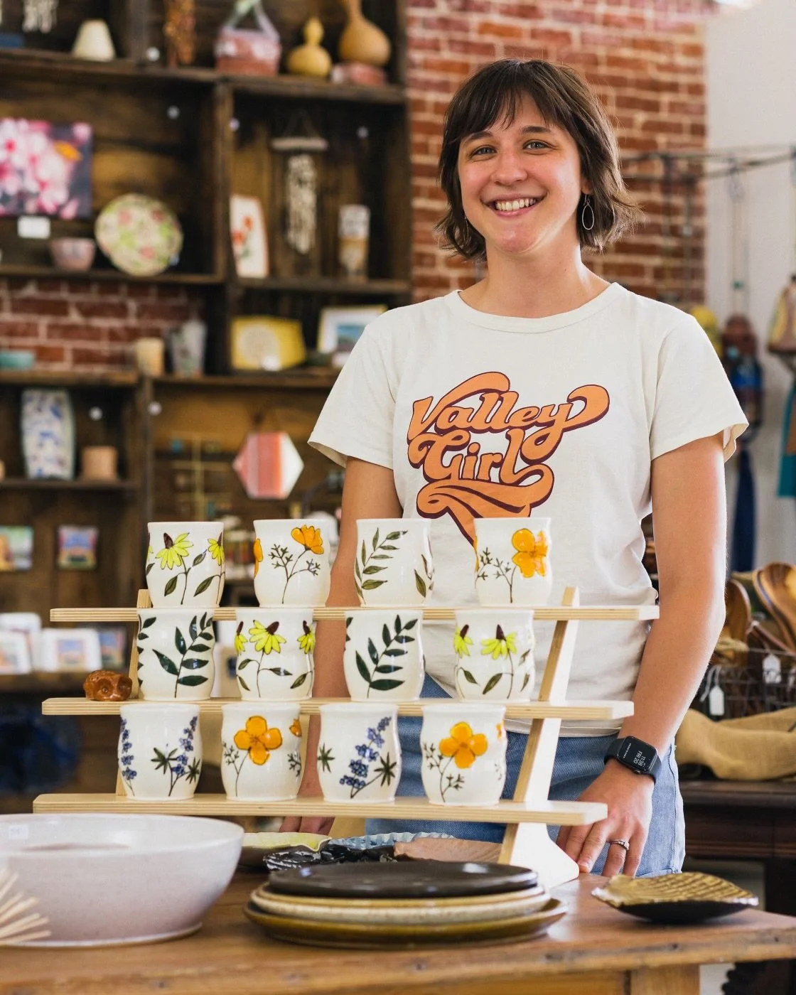 A woman smiling at a display of decorated ceramic cups with floral patterns at a craft fair or shop.