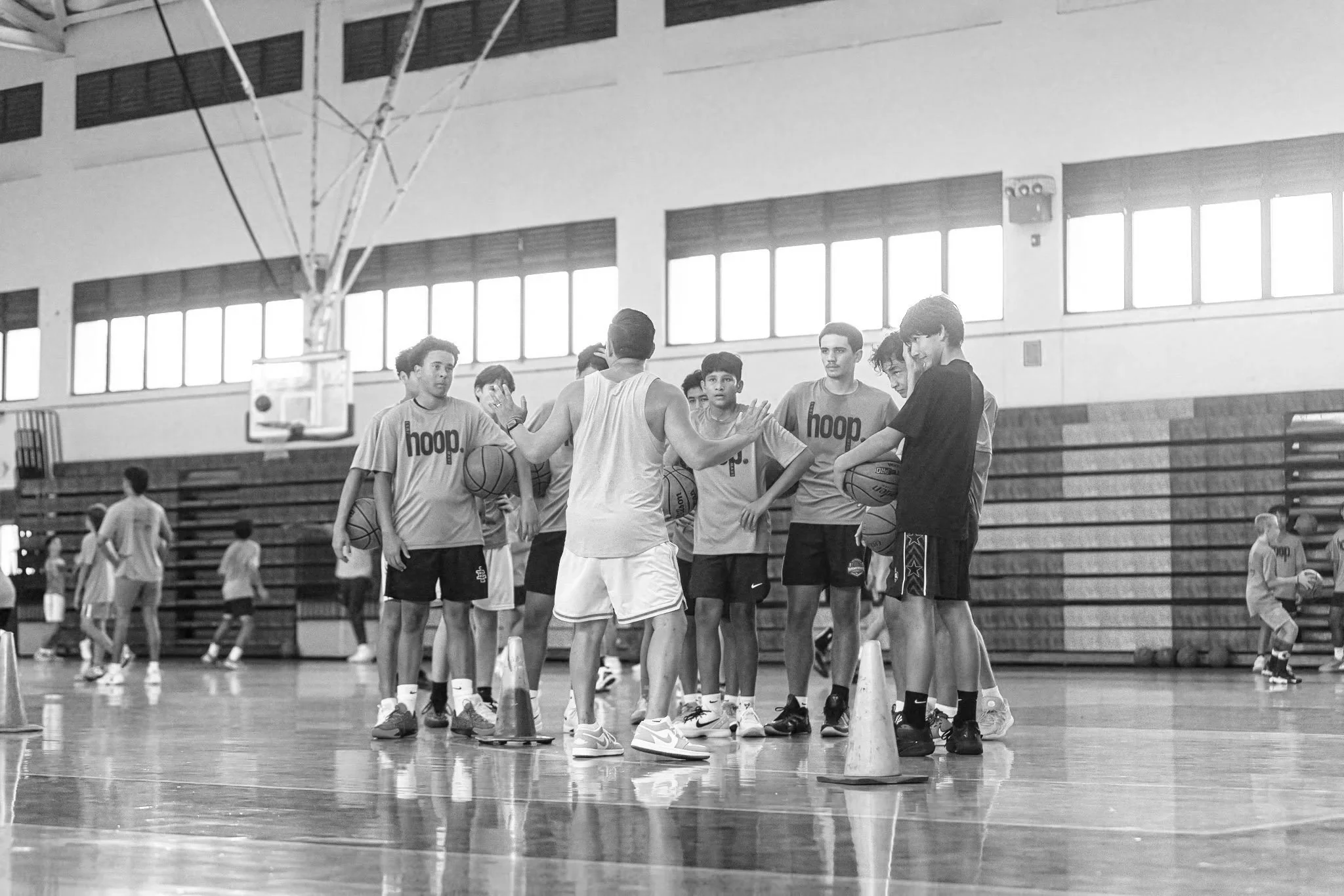 A basketball coach instructing a group of young players during practice in a gymnasium. The coach is talking and gesturing with his hands, while the players listen attentively, holding basketballs.