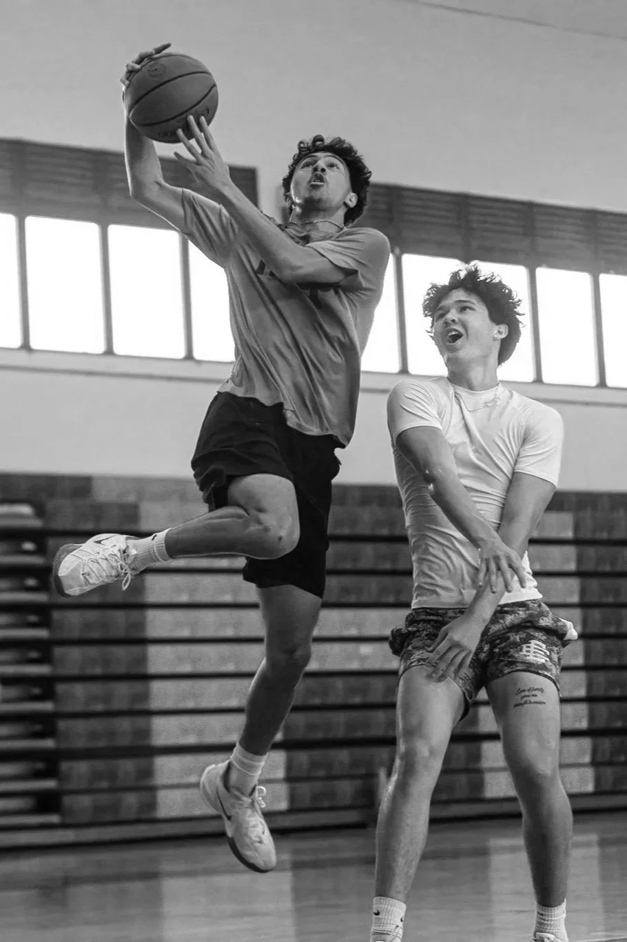 Two young men playing basketball indoors; one is jumping and reaching for the ball, the other is standing with an open mouth.