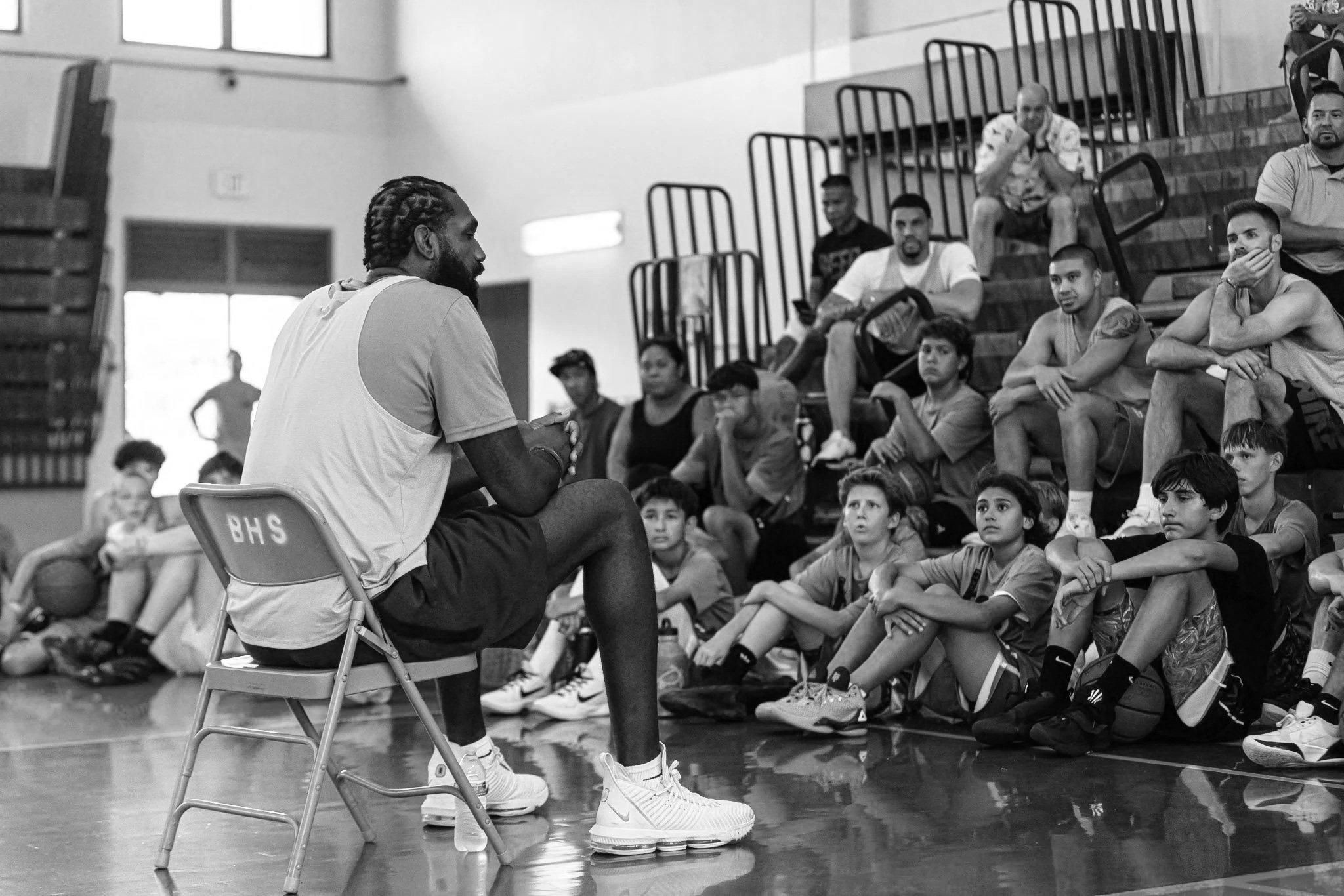 A man with braids in a sports jersey sits on a chair and speaks to a group of kids and teenagers seated on the floor and bleachers in a gymnasium.