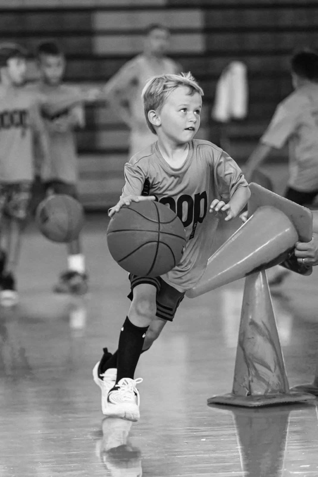 A young boy in sports attire dribbling a basketball during a practice drill in a gymnasium.