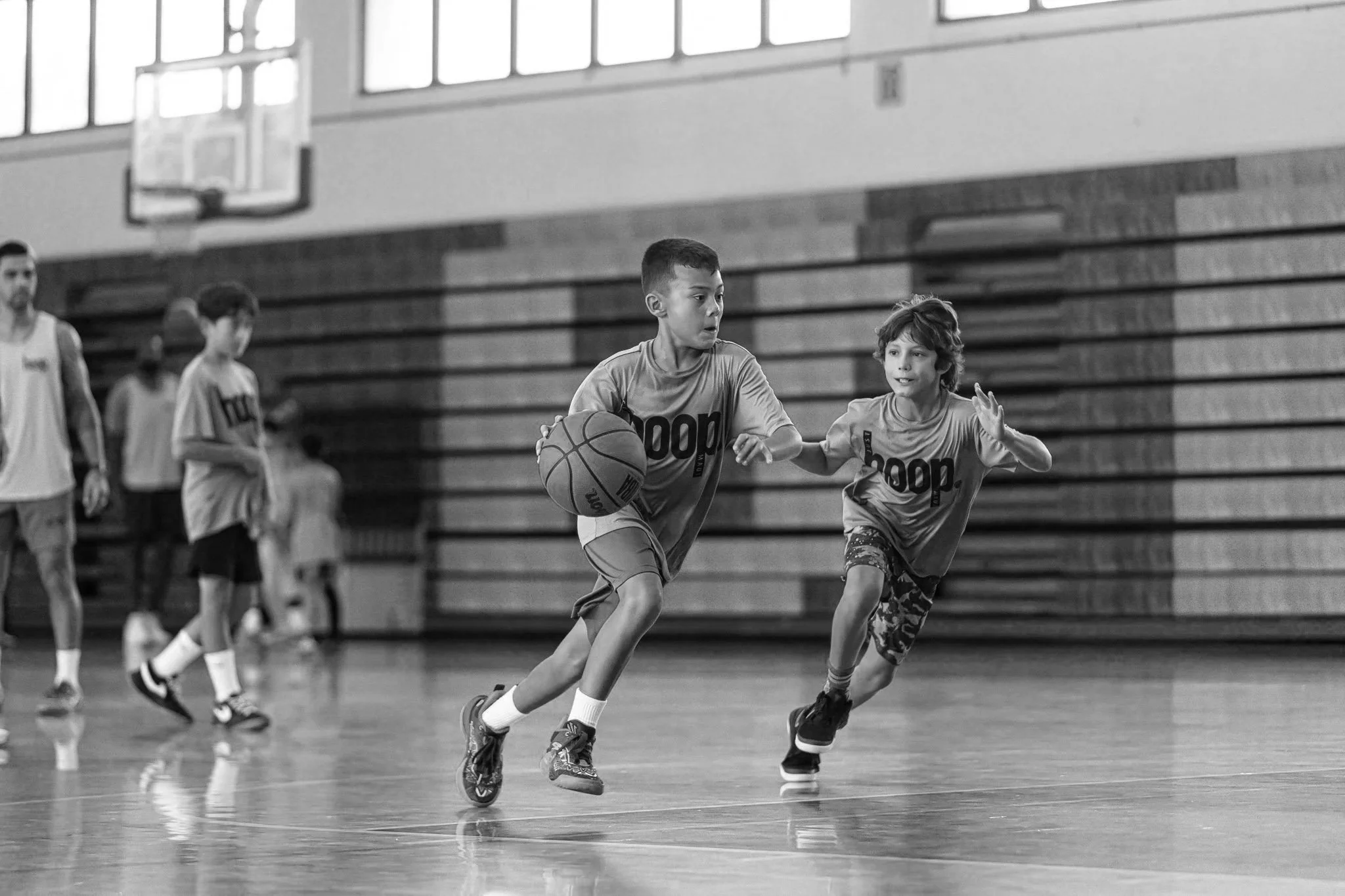 Two young boys playing basketball on an indoor court, with additional kids and a coach in the background.