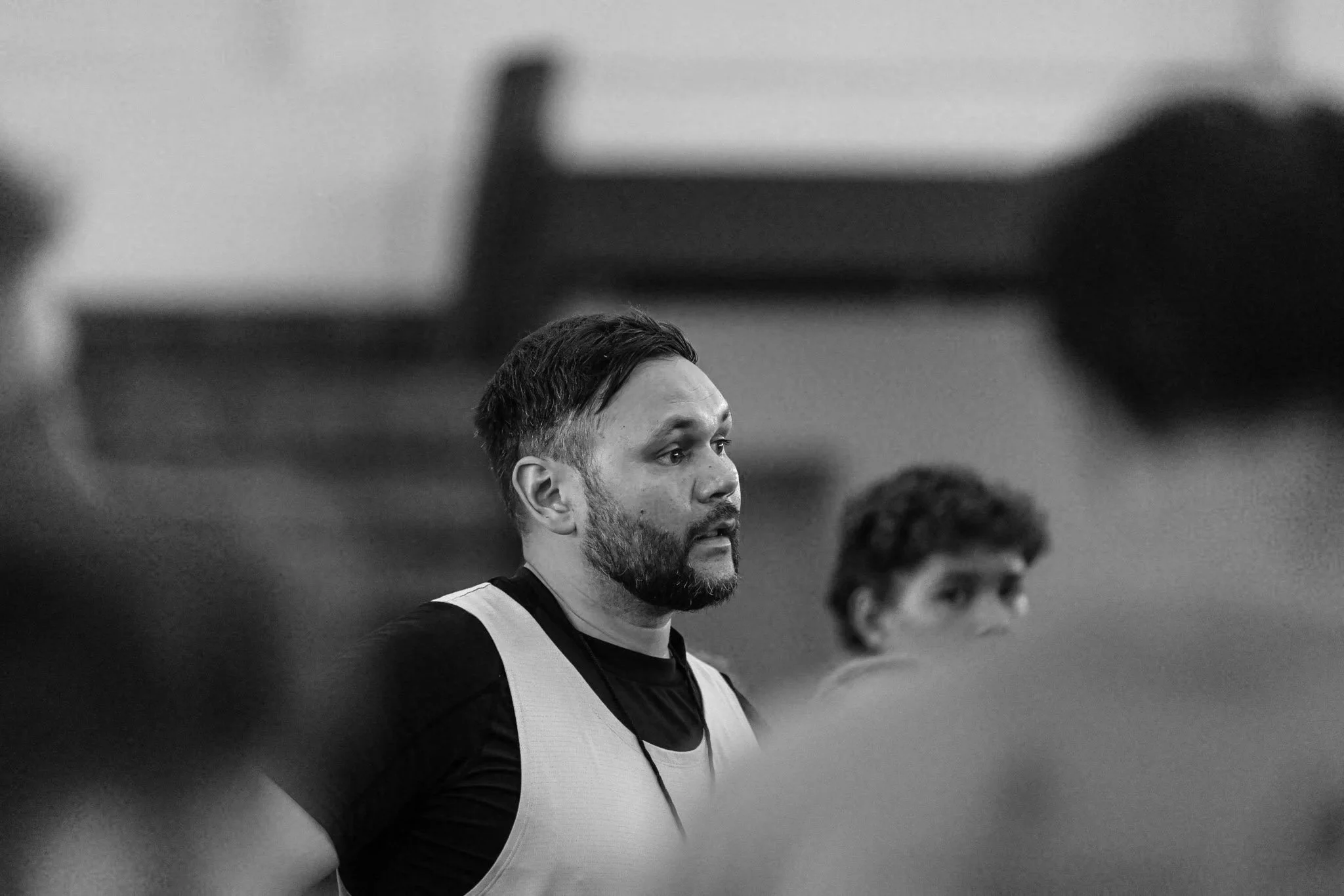 Black and white photo of a man with short dark hair, beard, and mustache, wearing a sports vest, looking straight ahead, with another person and blurred background.
