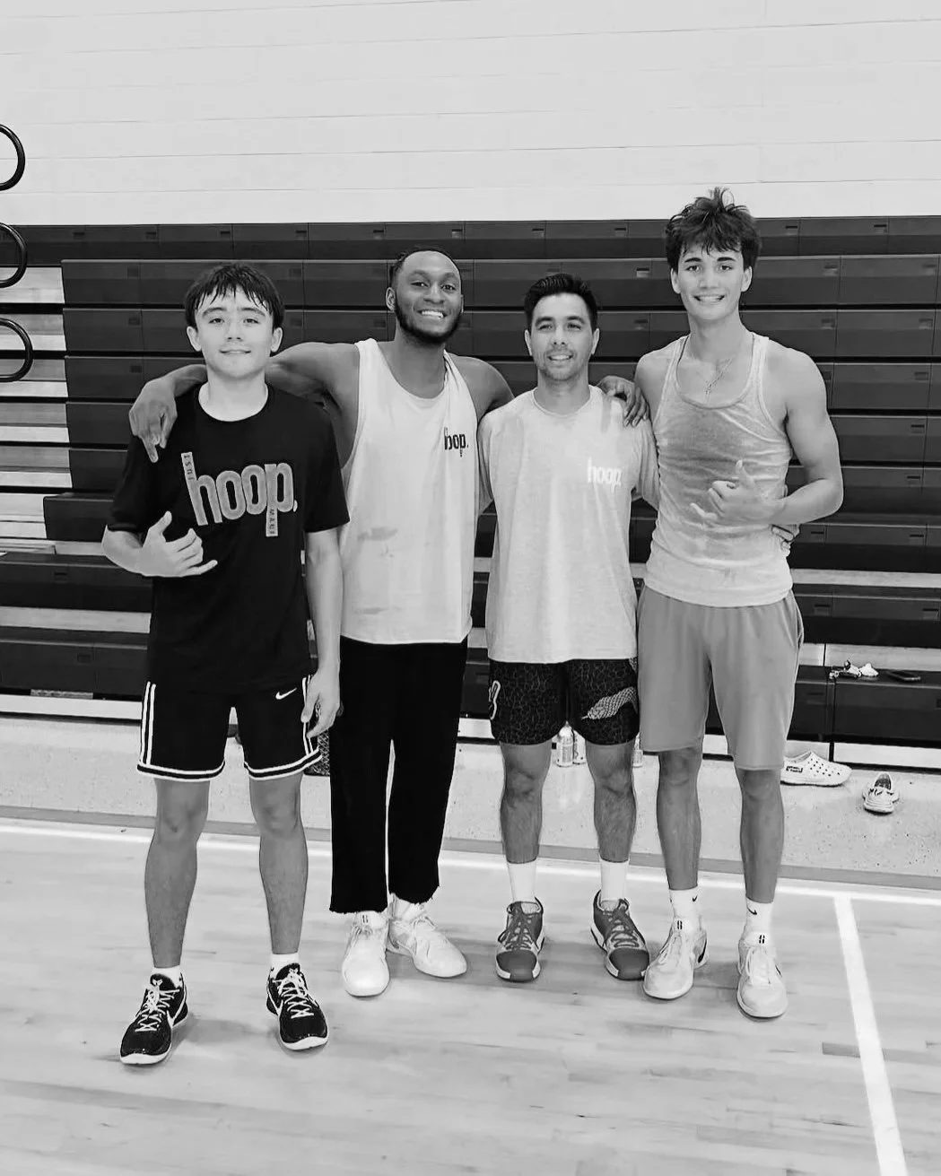 Four young men standing together in a gymnasium, three of them are wearing casual workout clothes, and two of them have 'hoop' branding on their shirts. They are smiling, with arms around each other, and standing on a basketball court.