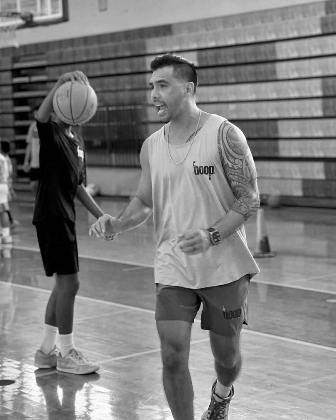 A man with tattoos on his arms wearing a sleeveless shirt and shorts, running on a basketball court, with a person holding a basketball in the background.