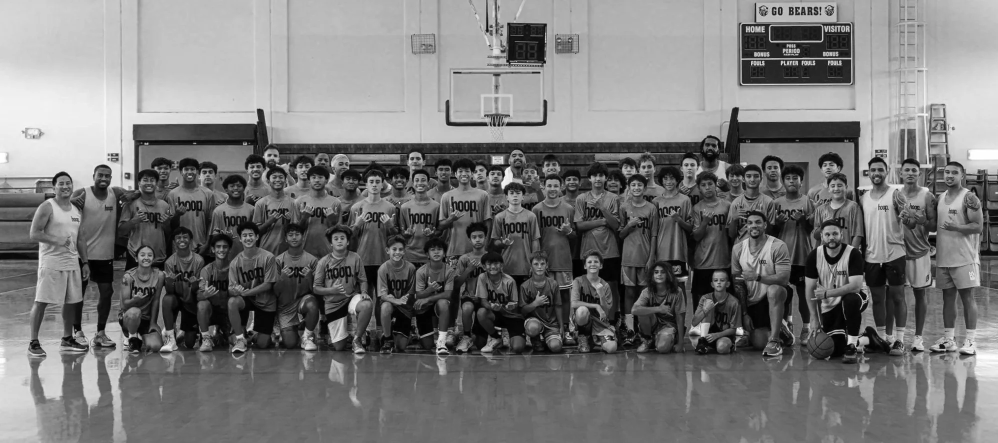 A large group of young basketball players and coaches posing for a team photo on an indoor basketball court. Some are kneeling in the front row, others standing behind. The players are wearing matching jerseys with the word 'hoop' printed on them. The scoreboard and basketball hoop are visible in the background.