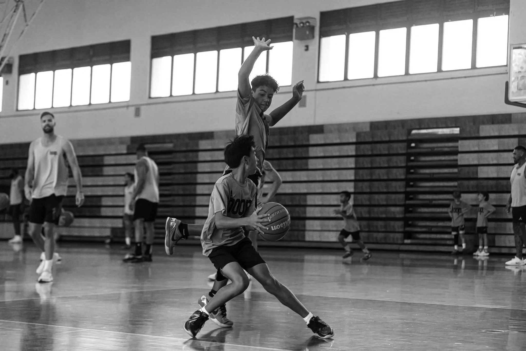 Young basketball player dribbling basketball while another player jumps to block him in indoor gym, with team members and coaches in background.