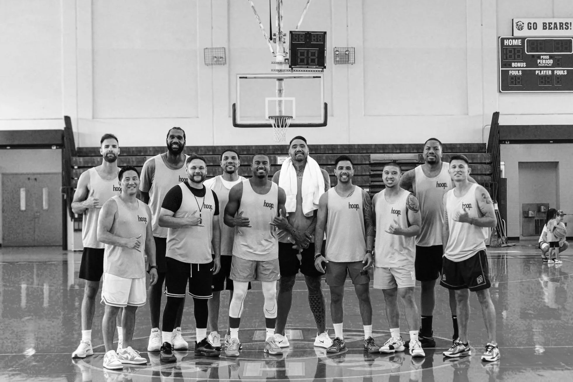 Black and white photo of a basketball team posing together on an indoor court, with a scoreboard and basketball hoop in the background.