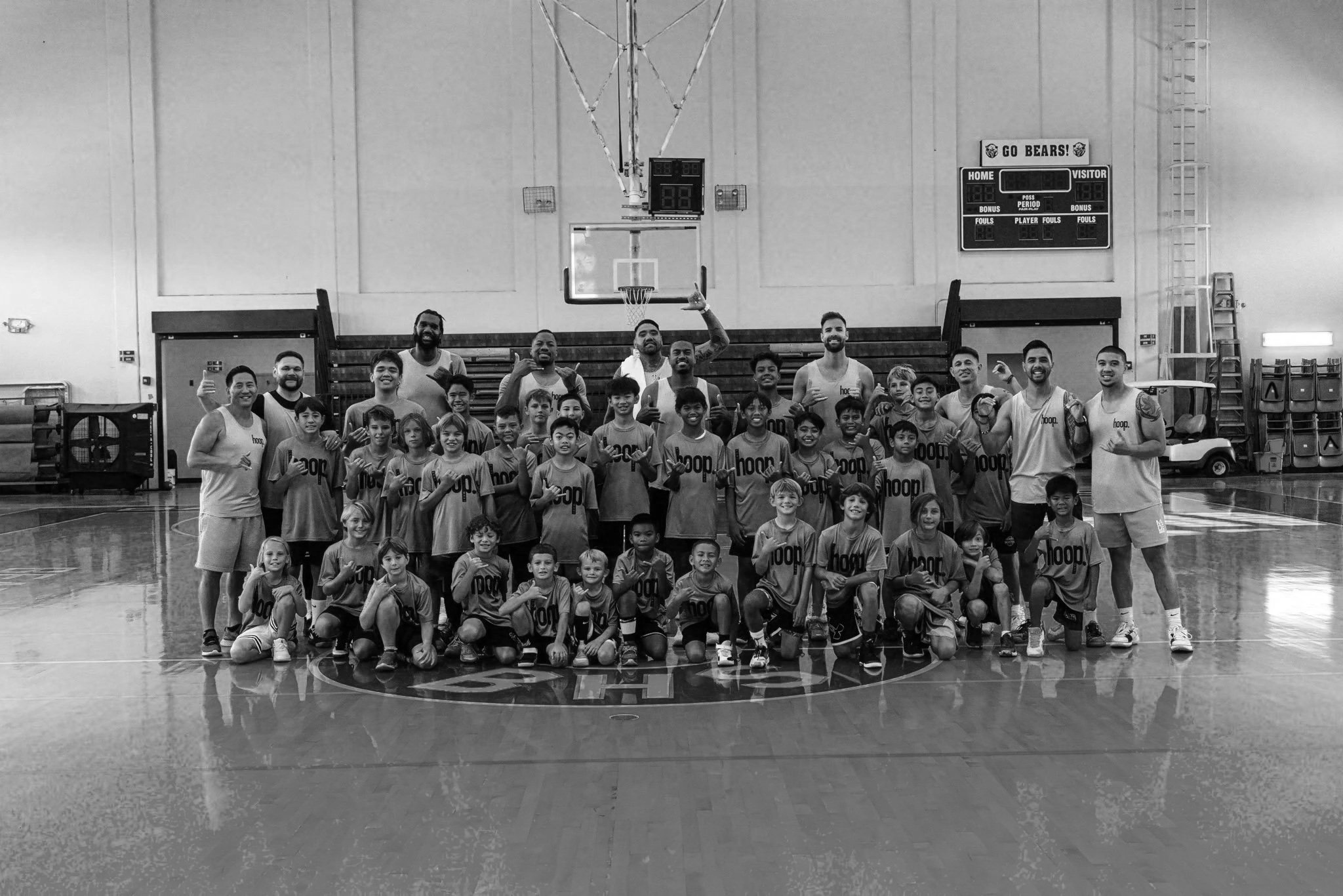 A large group of boys and coaches posing for a photo in a basketball gymnasium. The group is smiling and making various hand gestures. The gym has a basketball hoop, scoreboard, and gym equipment in the background.