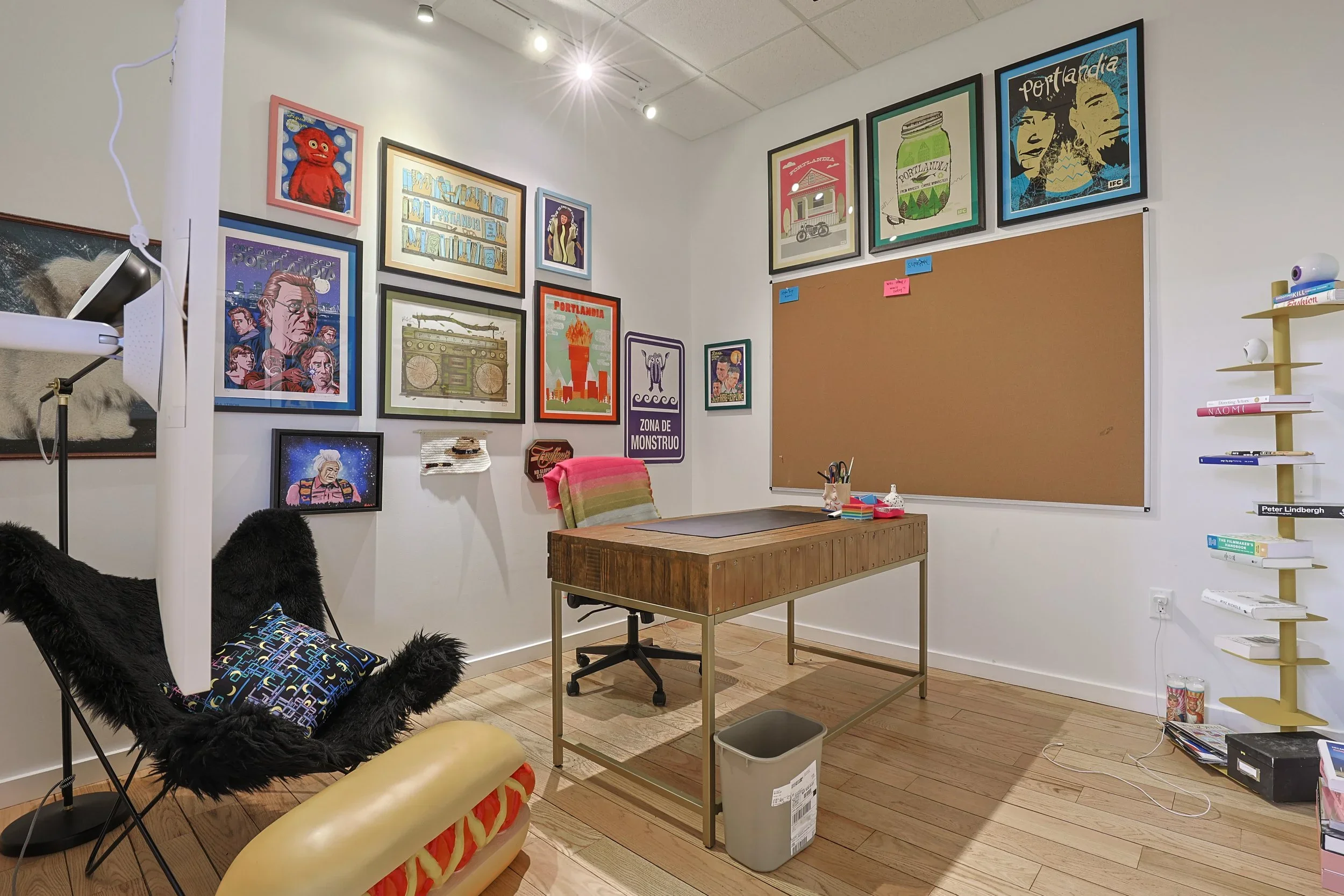 An art studio or gallery space with various framed artwork on the white walls. There is a wooden desk with art supplies, a chair with a colorful blanket, and a black furry chair with a pillow. A corkboard is on one wall, and a shelving unit with book