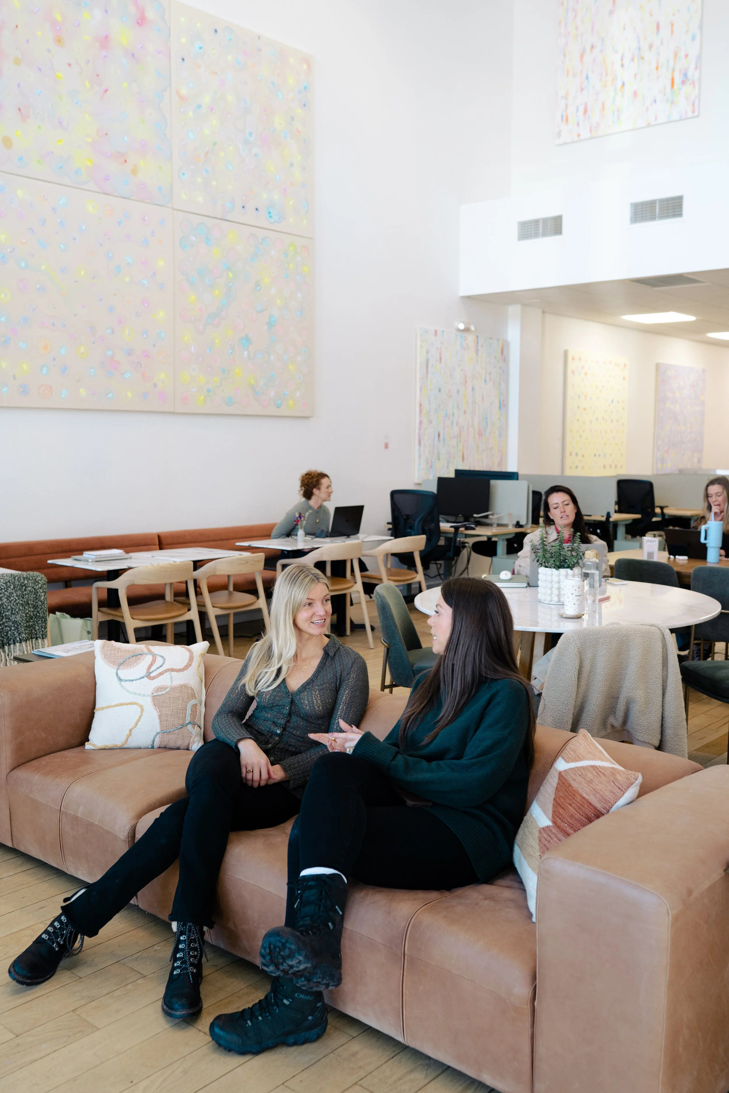 Two women sitting on a tan leather sofa with decorative pillows talking to each other, in a bright modern office or coworking space with large abstract colorful paintings on the white walls and other people working in the background.