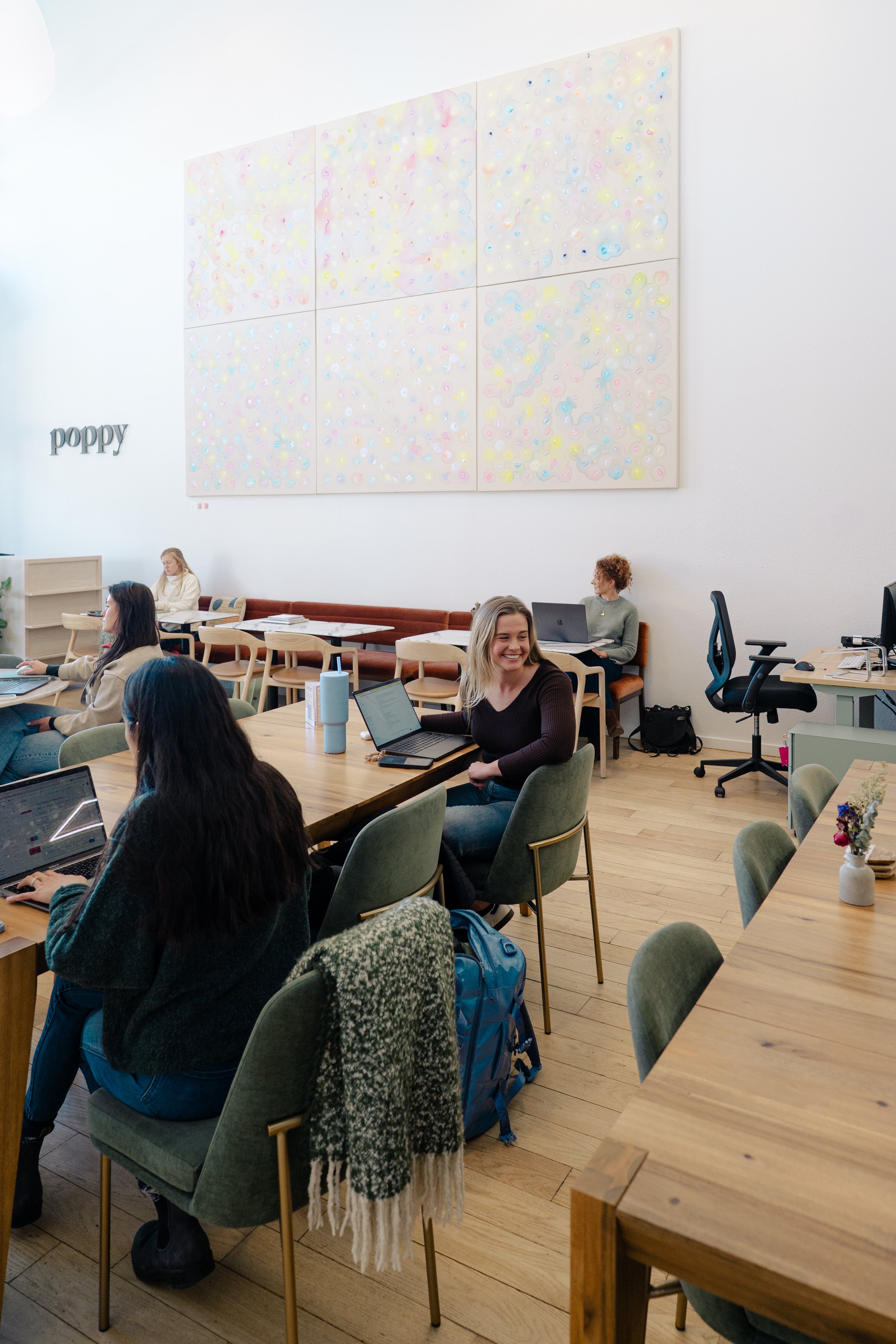 A group of people working at a co-working space or office, with some using laptops at a wooden table and others sitting on a long cushioned bench. There is a large white wall with a colorful, abstract artwork and the word "poppy" in lowercase letters.