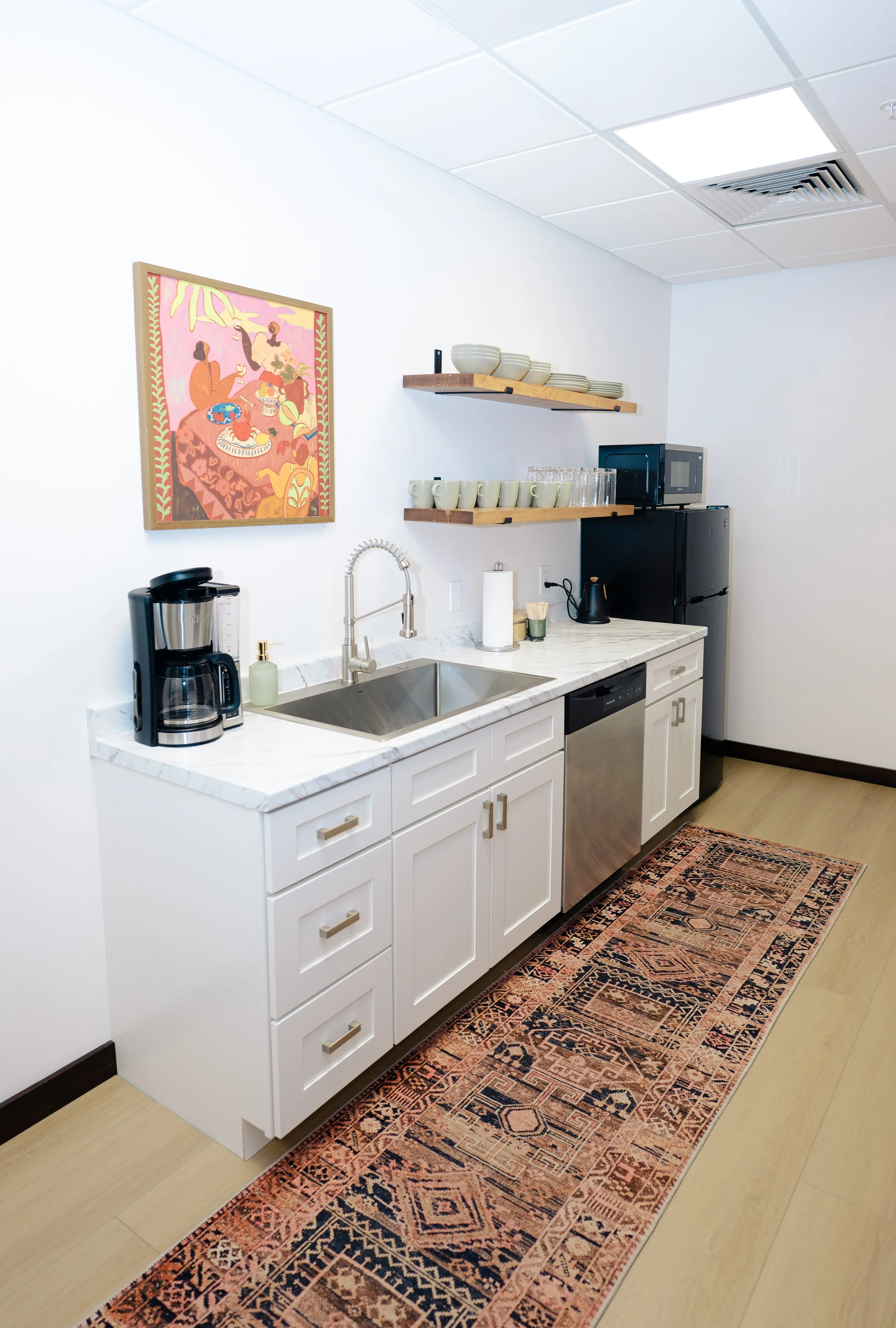 A modern kitchen with an island countertop, white cabinets, and a dark refrigerator. Open shelves hold bowls, cups, and glasses. There is a coffee maker and a soap dispenser on the counter. A colorful, patterned rug runs along the wooden floor.