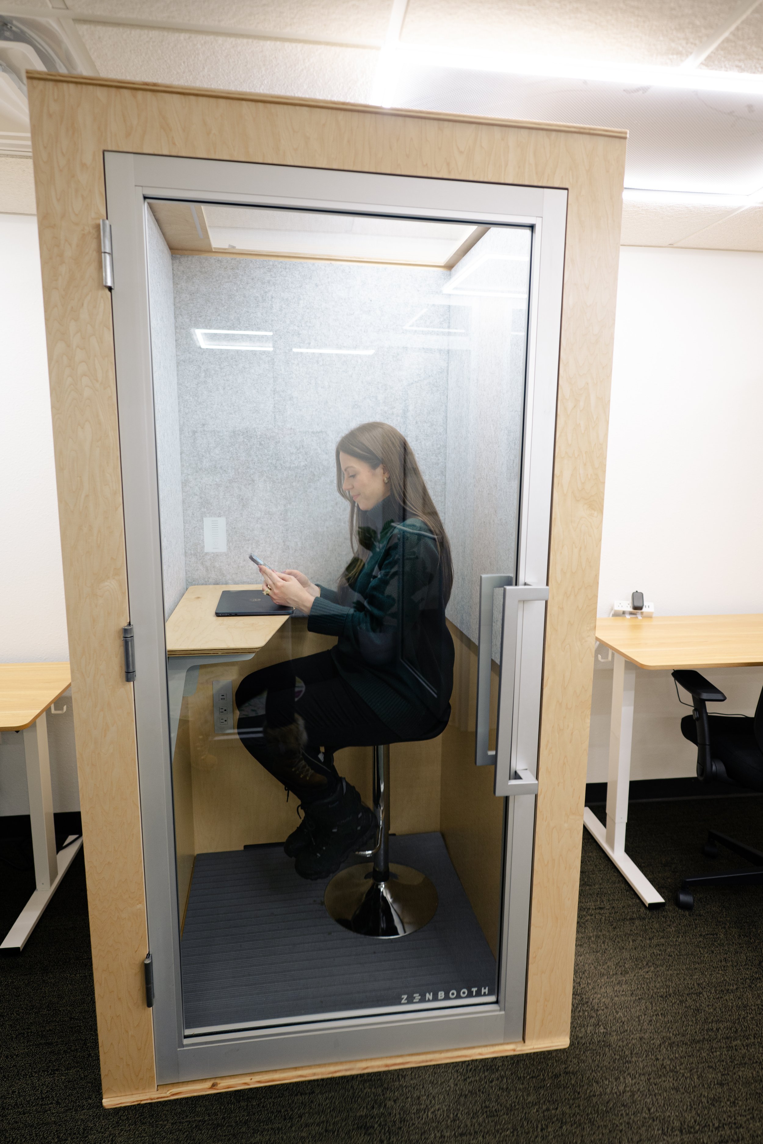 Woman sitting in a soundproof booth at her desk, using her phone, inside an office.