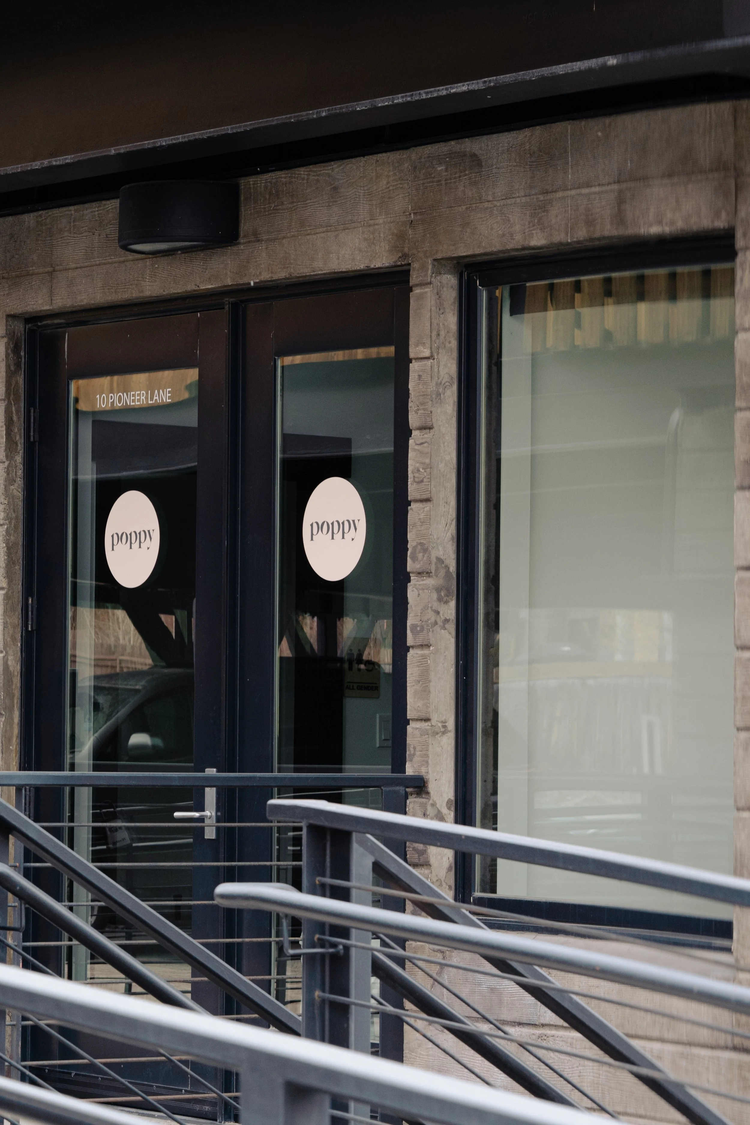 Close-up of exterior entrance to a building with glass double doors, a black door handle, a window on the right, a sign with the number 10Pioneer Lane, and a shop sign that says "poppy". There is a black speaker mounted above the doorway and black metal railings on a staircase leading to the entrance.