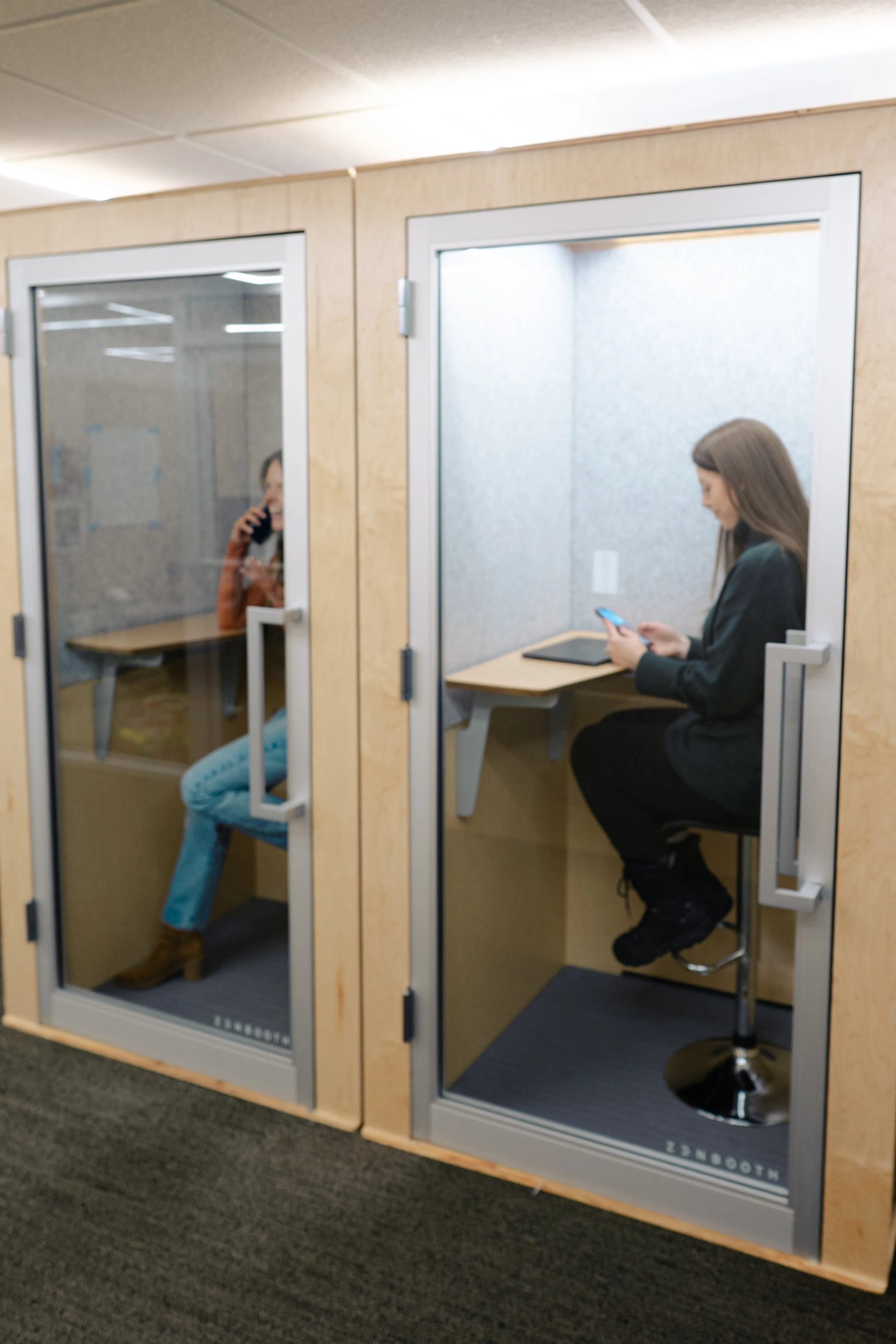 Two women sitting inside soundproof booths, one using a phone and the other on a call, in an office or co-working space.