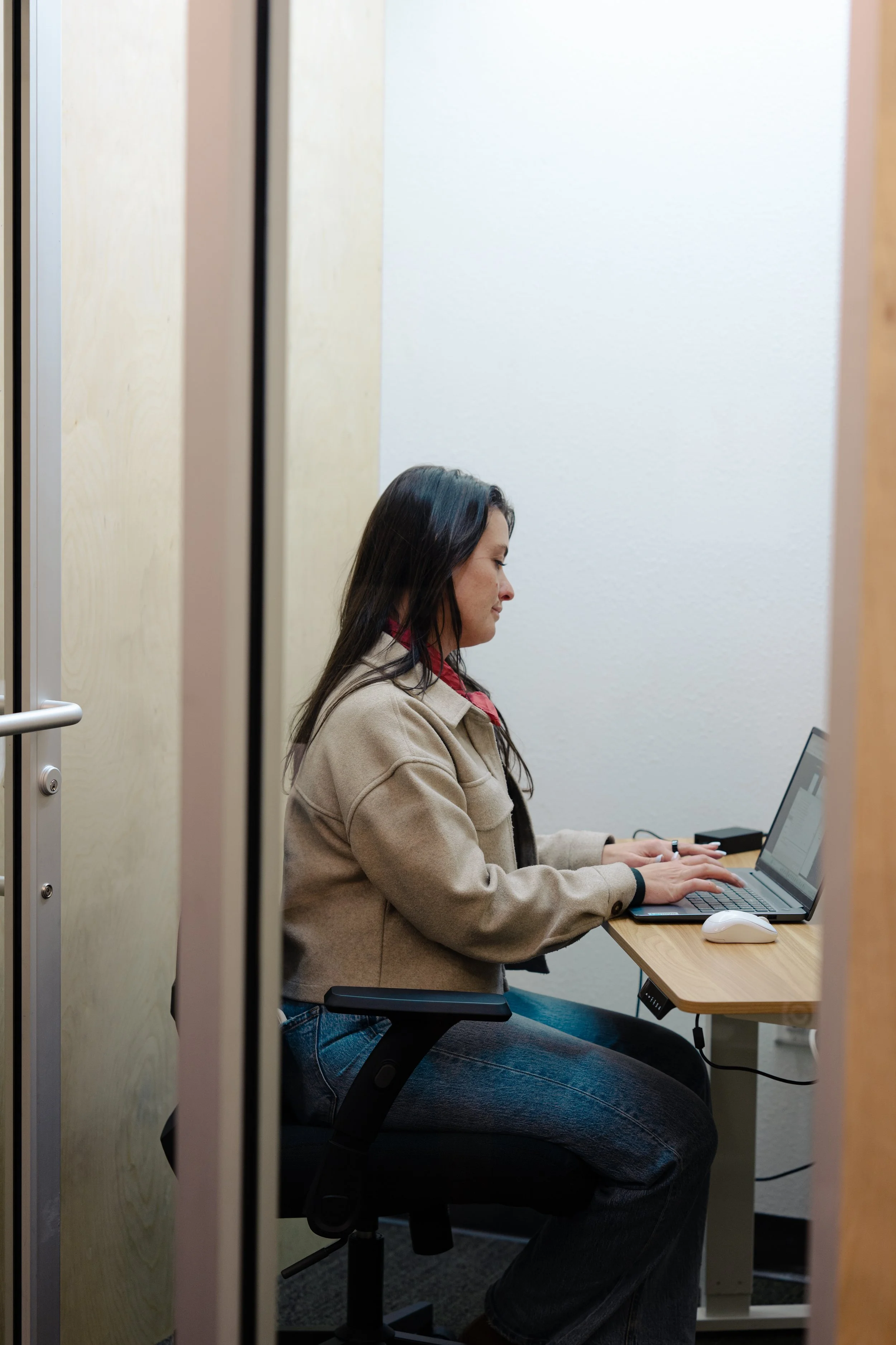 A woman sitting at a desk in an office, using a laptop with a computer mouse beside her.