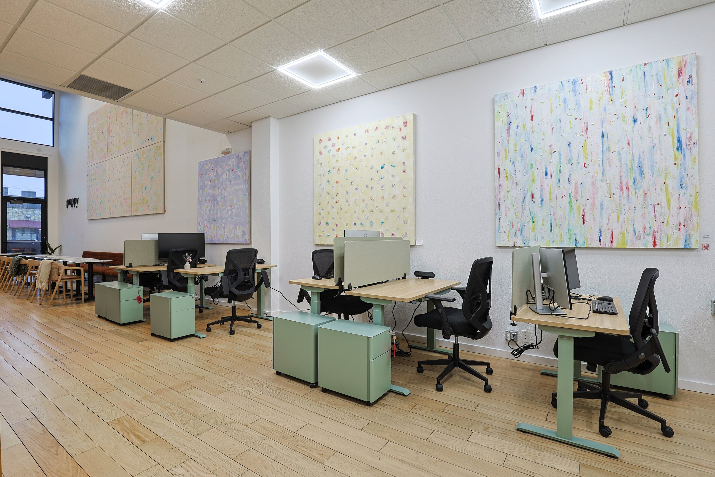 Office space with desks, chairs, computers, and abstract paintings on the white walls. Large window on the left side, wooden flooring, ceiling lights.