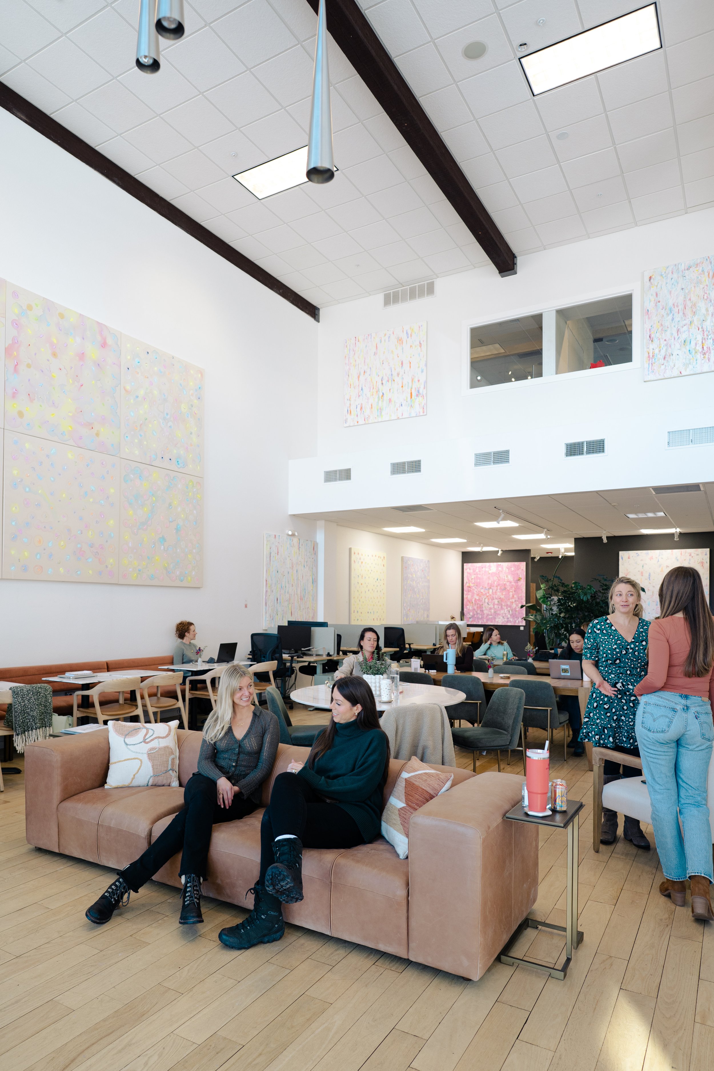 A modern, brightly lit office or coworking space with art on the walls, people working on laptops, and everyone socializing. There are two women sitting on a tan couch in the foreground having a conversation, and a group of people behind them working or talking. The room has light wood flooring, a high ceiling with hanging lights, and a lot of natural light coming through large windows.