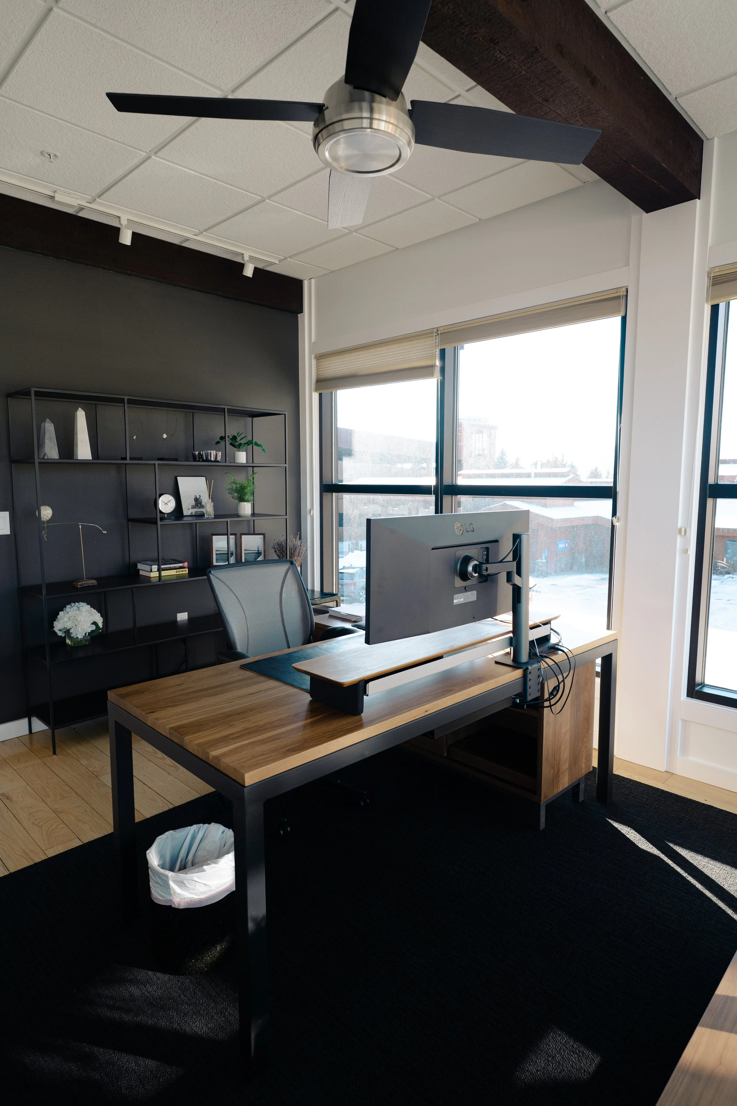 Modern office with a wooden desk, ergonomic chair, computer monitor, black bookshelf, large windows with blinds, ceiling fan, and a black rug with a white trash bin.