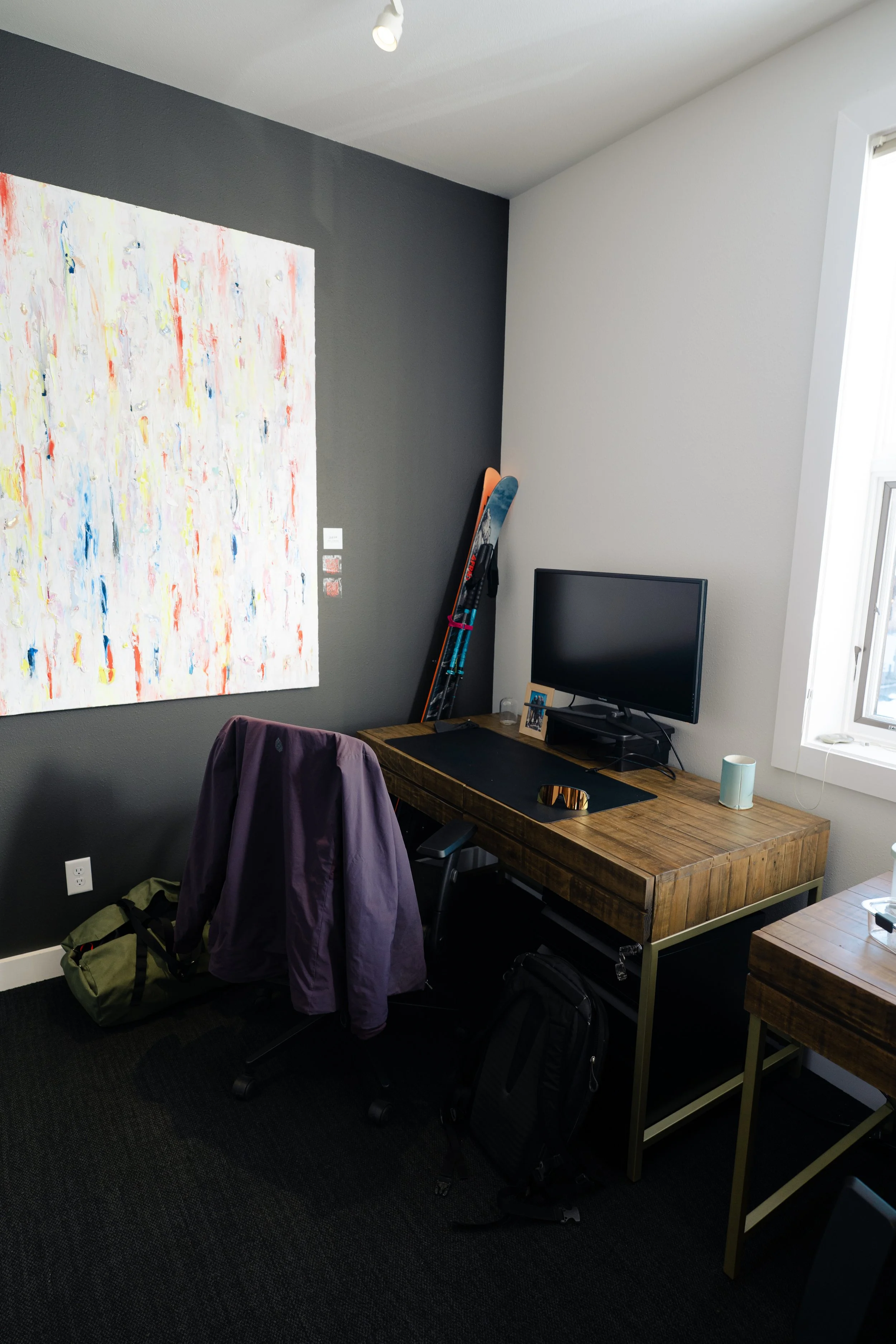 A workspace with a wooden desk, computer monitor, a purple jacket hanging on a chair, skis leaning against a gray and white wall, an abstract painting, and various personal items including a backpack on the floor and a cup on the desk.
