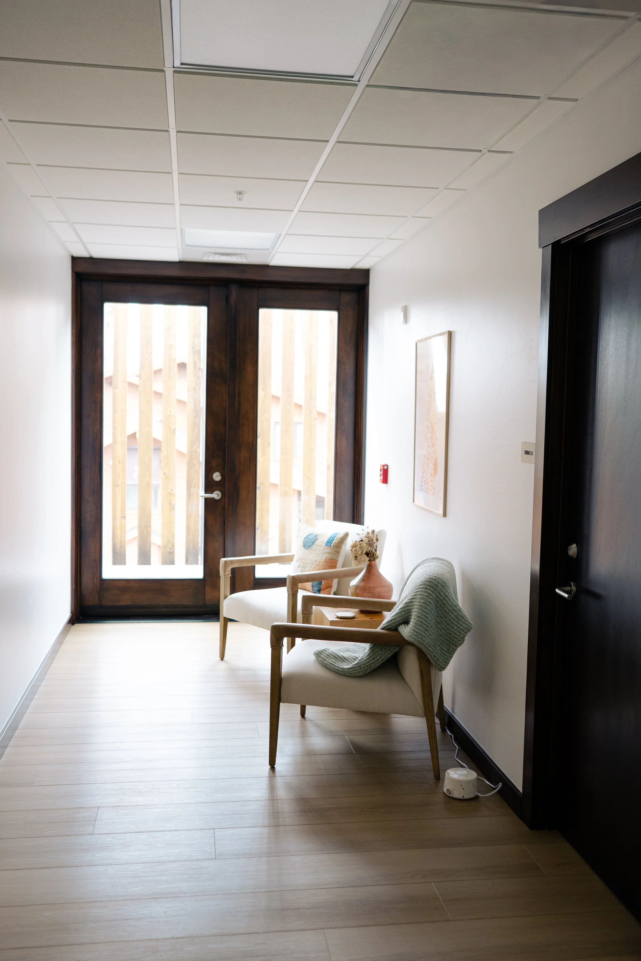 Interior view of a small waiting area with two chairs, a small wooden side table with a vase, and framed artwork on the wall. There is a large glass door with wooden framing leading outside, with vertical wooden slats visible through the glass. The ceiling has white tiles and there is a fire alarm on the wall.