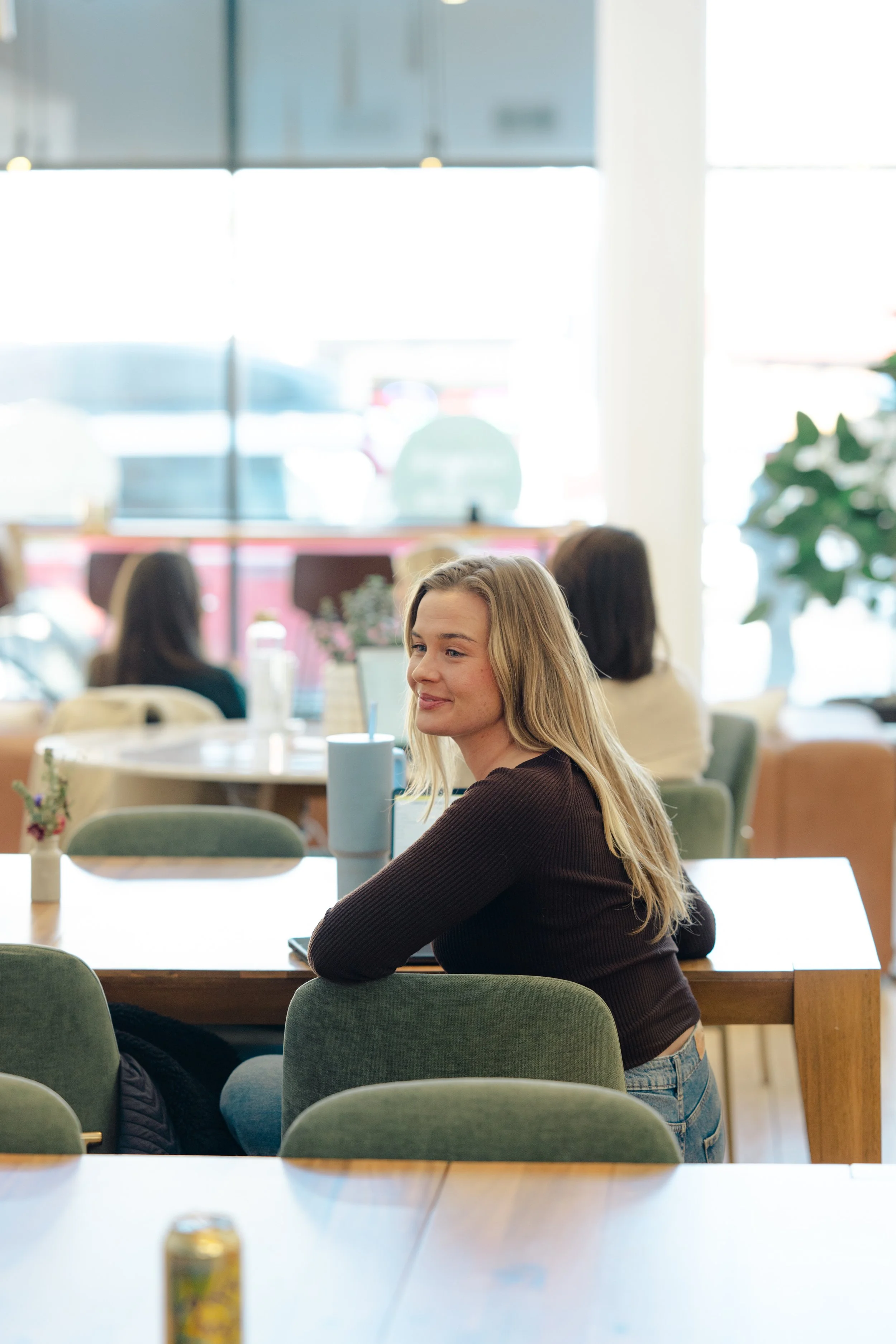 A young woman with long blonde hair, wearing a dark sweater and jeans, sits at a table in a bright cafe. She is smiling slightly and looking off to the side, with a large window behind her letting in natural light. Other people are in the background, seated at tables, and there are potted plants and decorative items in the cafe.