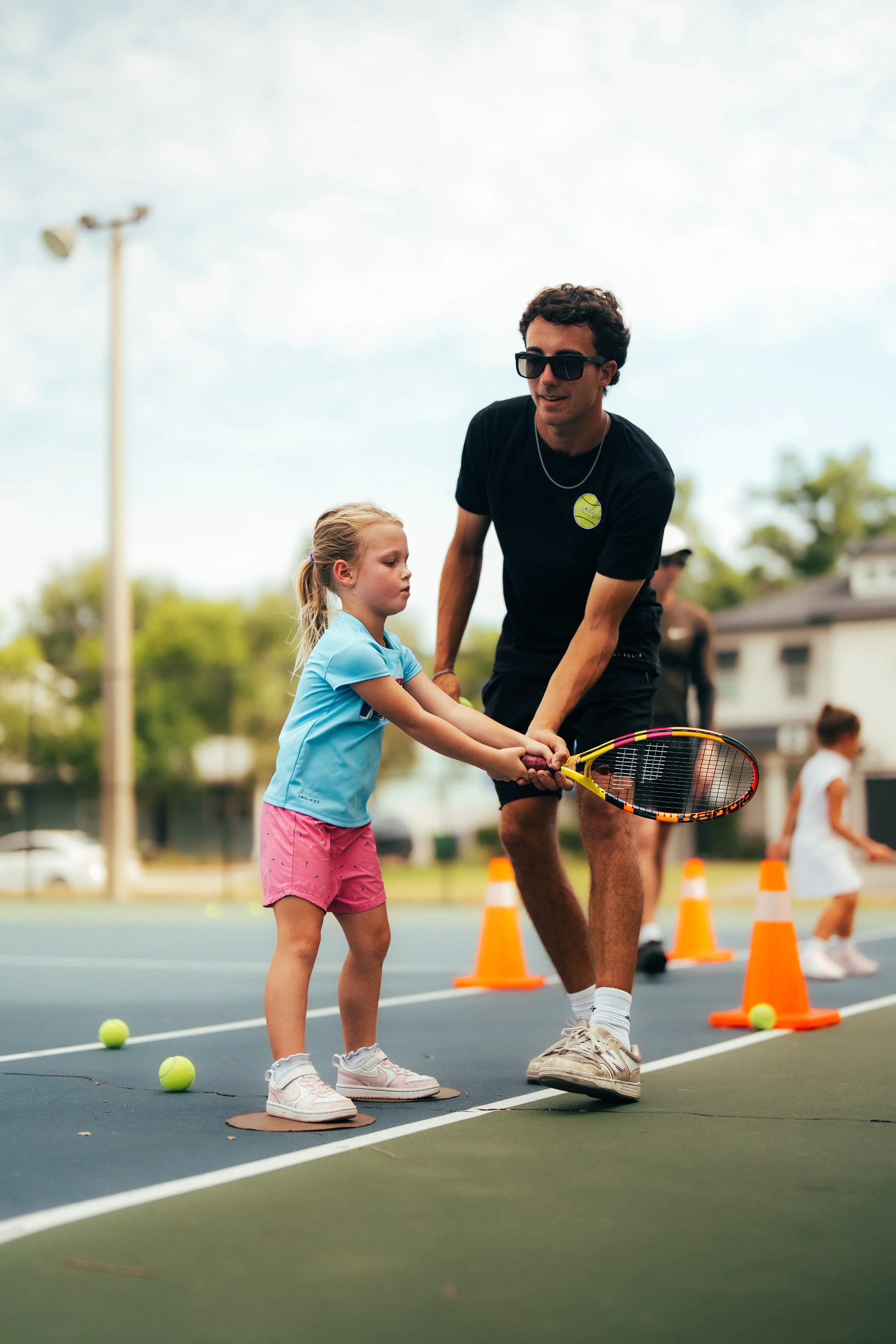 A tennis coach teaching young girls playing tennis on a blue court outdoors, with trees and a fence in the background.