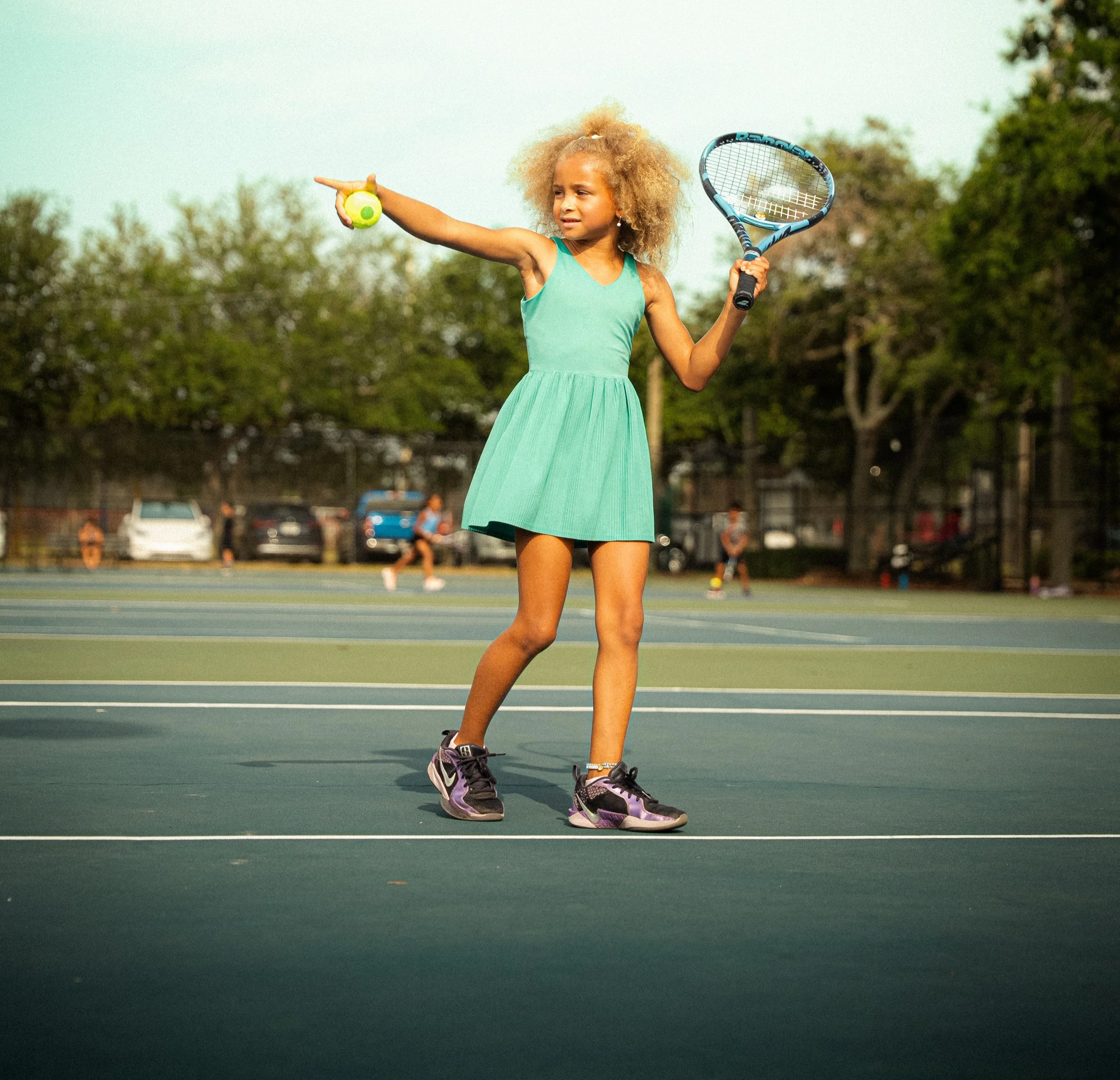 Young girl on tennis court holding tennis racket and tennis ball, pointing to her left.