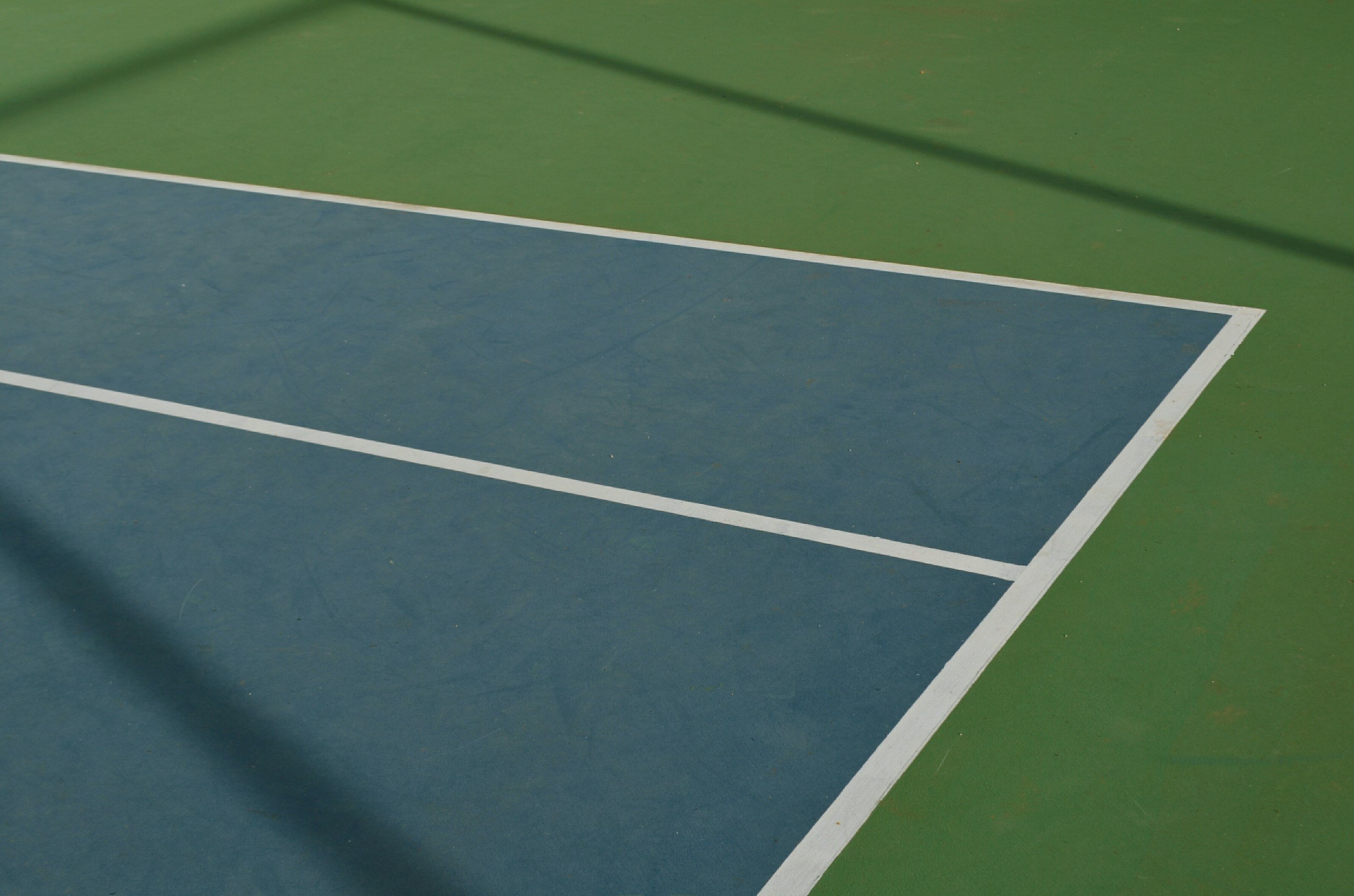 A section of a tennis court with a blue playing area and green surroundings, marked with white lines.