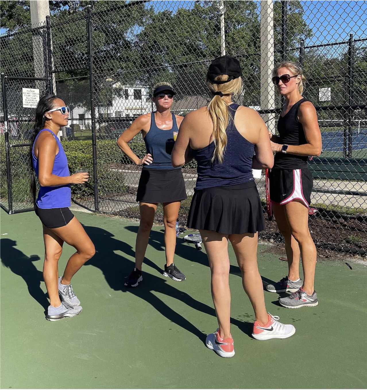 Four women in athletic wear standing and talking on a tennis court during the day.