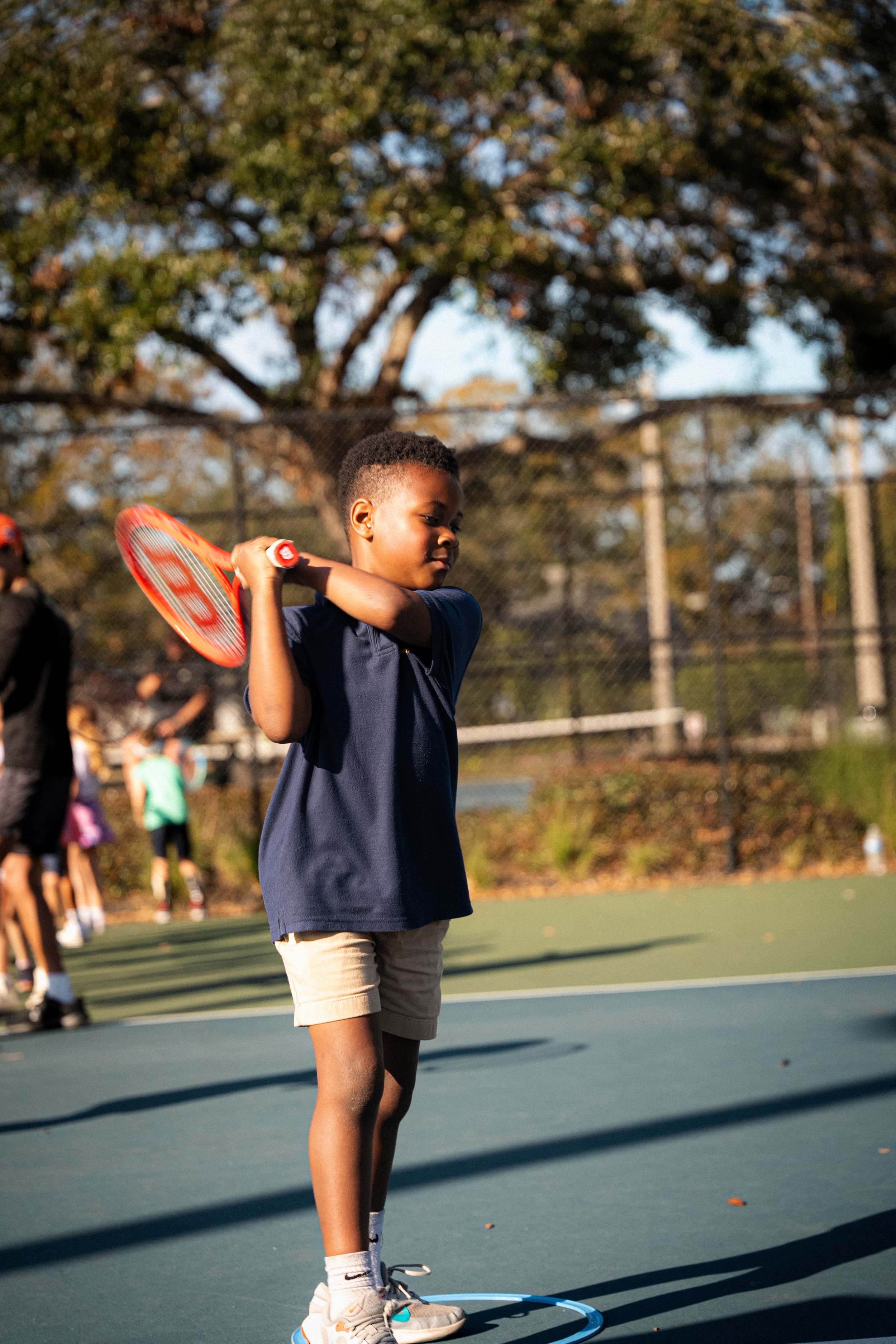 A young boy playing tennis on an outdoor court, holding a tennis racket and preparing to hit the ball, with other children in the background.