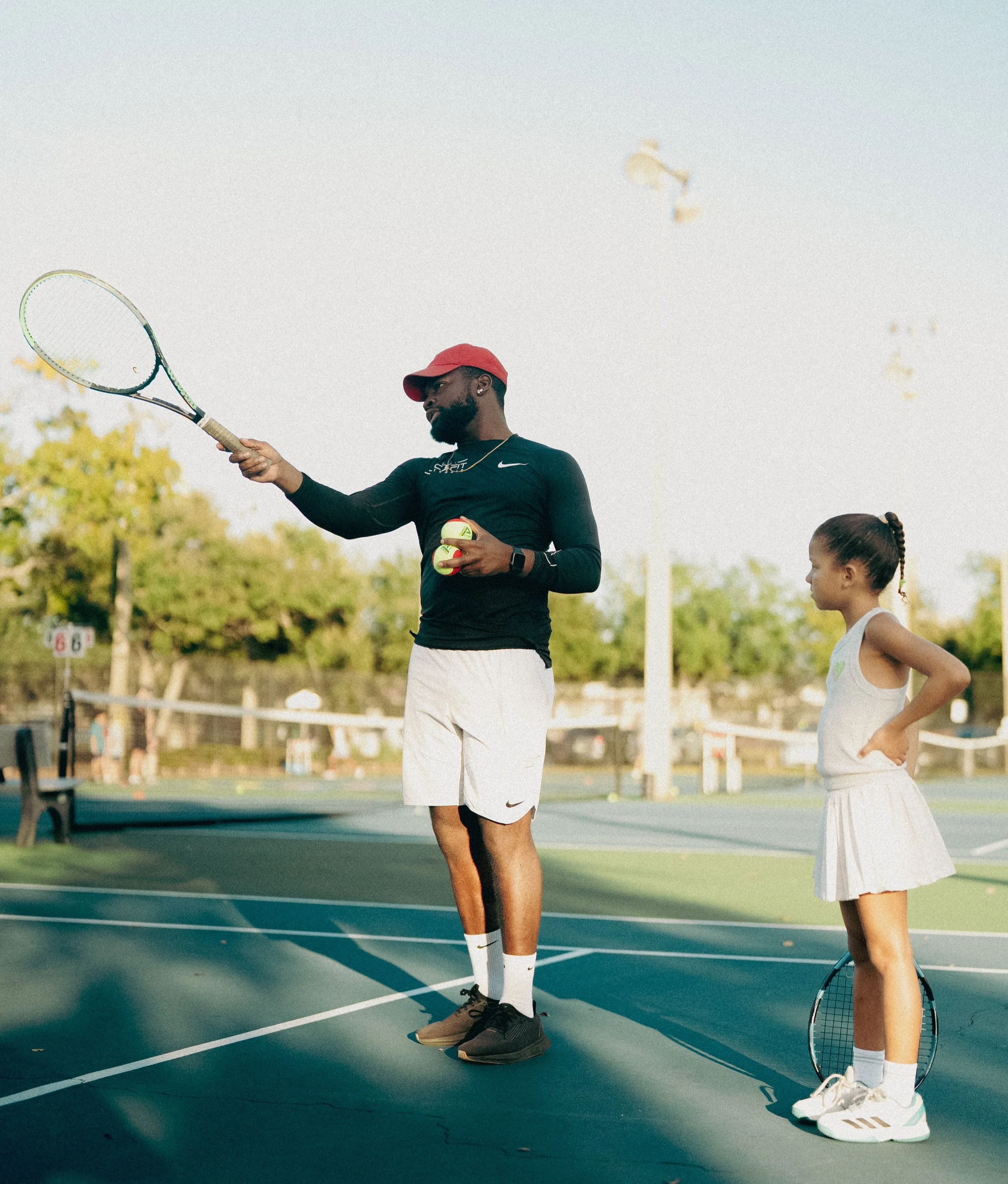 A man and a young girl on an outdoor tennis court. The man is holding tennis balls and a racket, wearing a black long sleeve shirt, white shorts, and a red cap. The girl is standing with her hands on her hips, holding a tennis racket, wearing a white tank top and skirt, with her hair tied back.
