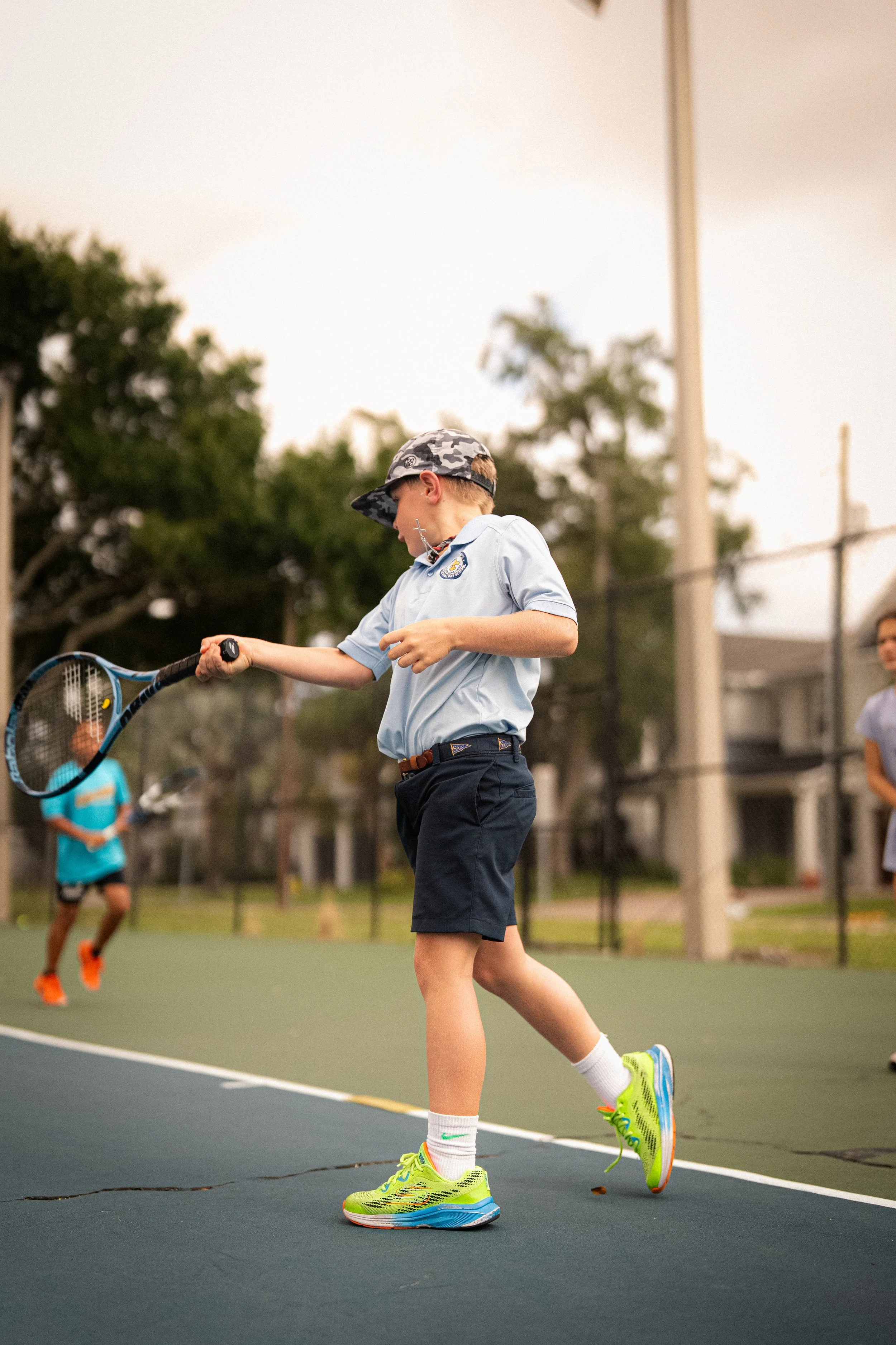 A boy playing tennis on a court with a girl and a boy in the background, trees, and a sky with clouds.