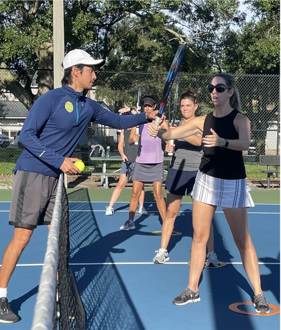 A tennis coach is instructing a female player during a practice session, with other women standing behind them on the tennis court.