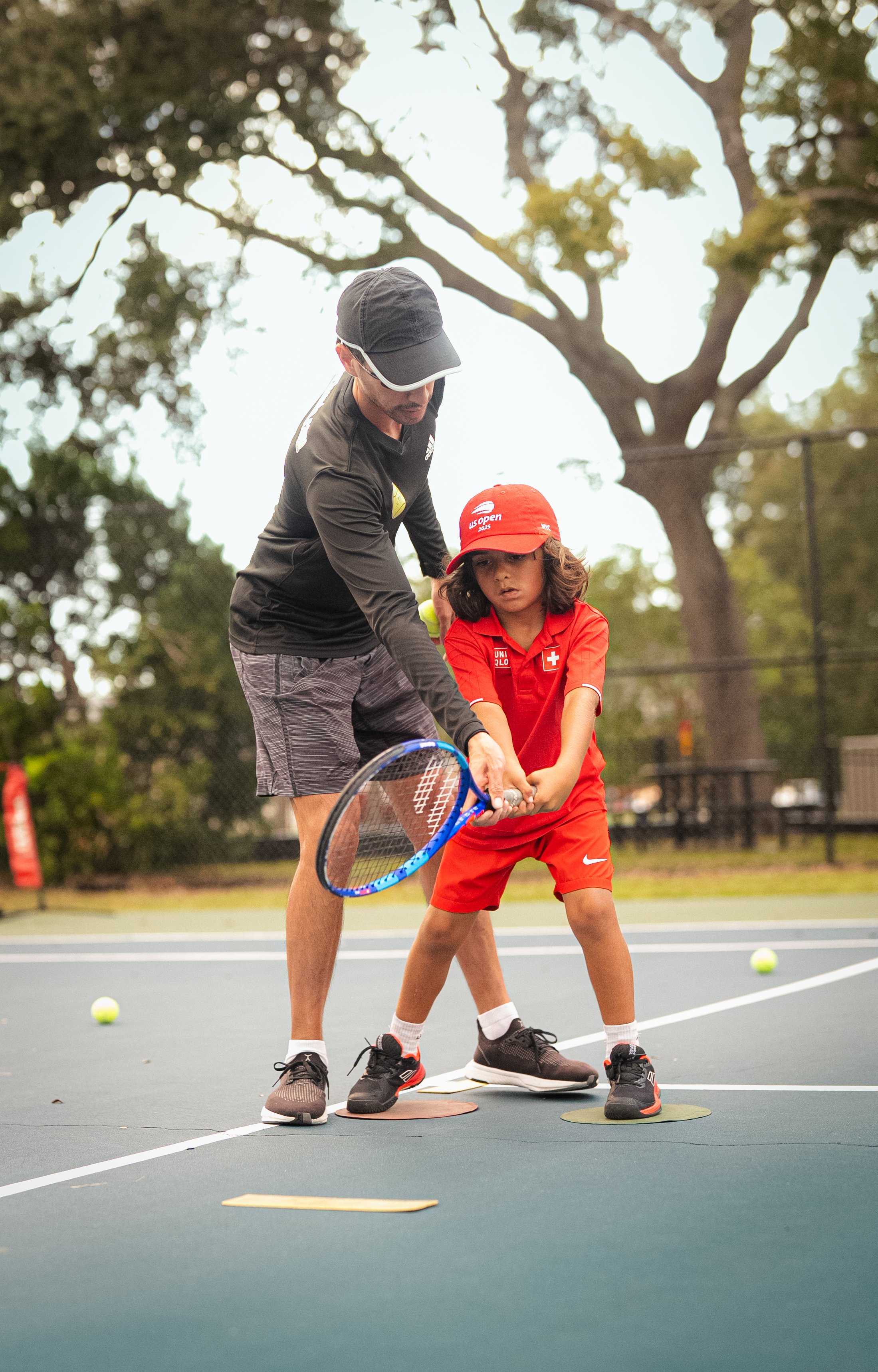 A coach and a young girl on a tennis court practicing tennis. The girl is wearing a red tennis outfit and a red cap, while the coach is dressed in dark sportswear. Several tennis balls and court markers are visible on the ground.