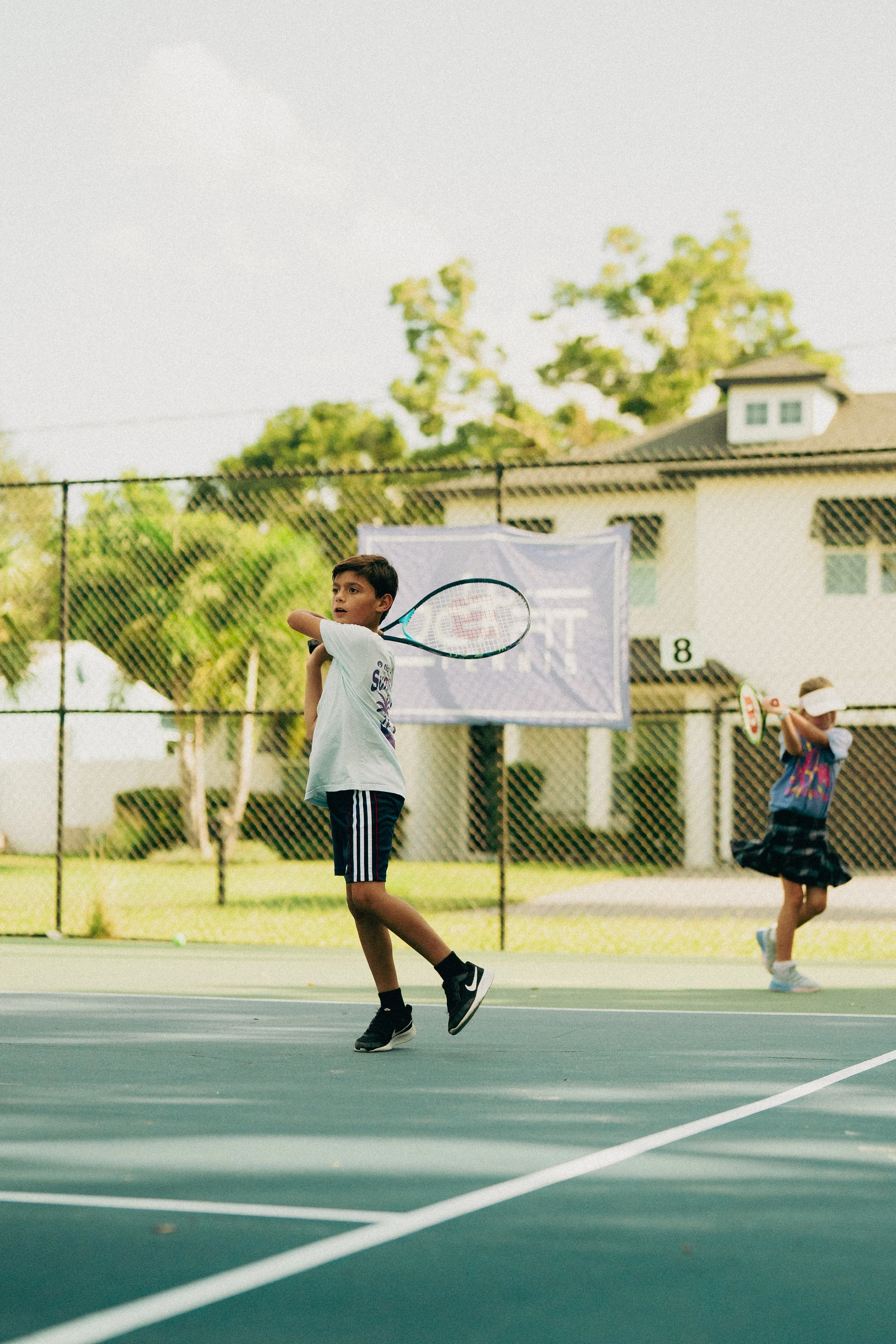 A young boy playing tennis on an outdoor court, holding a tennis racket and preparing to hit the ball, with other children in the background.