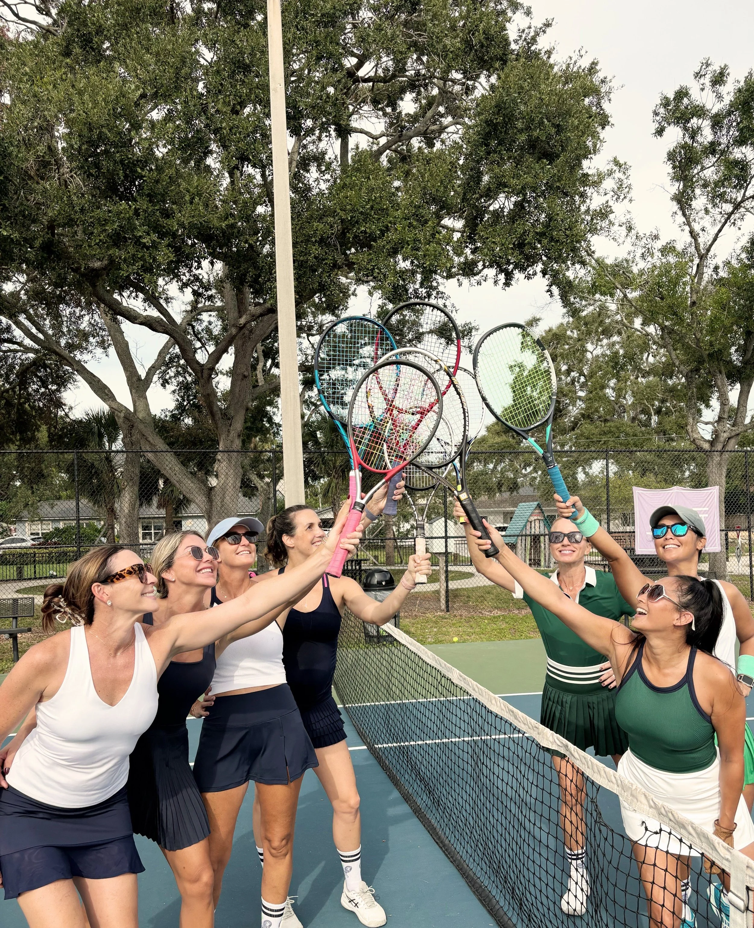 Group of women on a tennis court, smiling and holding tennis rackets in the air, celebrating.
