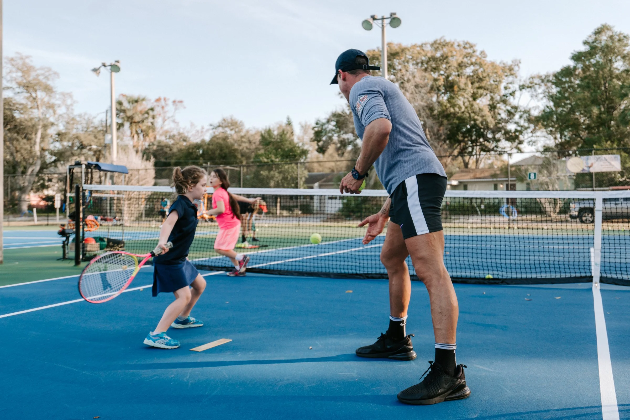 A tennis coach teaching young girls playing tennis on a blue court outdoors, with trees and a fence in the background.