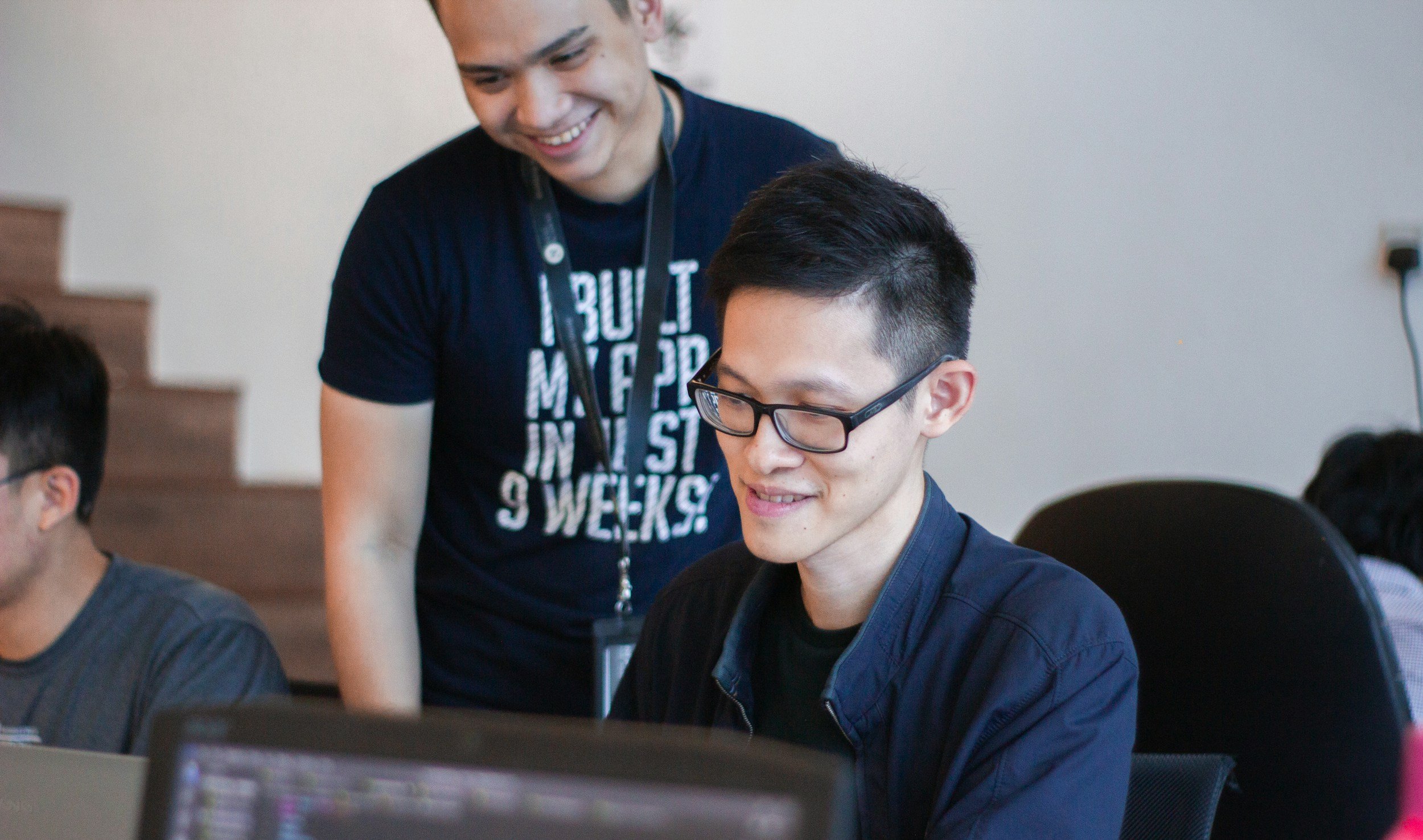 Two young men, one wearing glasses, working together at a table with laptops in a classroom or meeting room. A third person is partially visible on the left. The man standing is smiling and wearing a T-shirt with text, leaning over to assist the seated man.