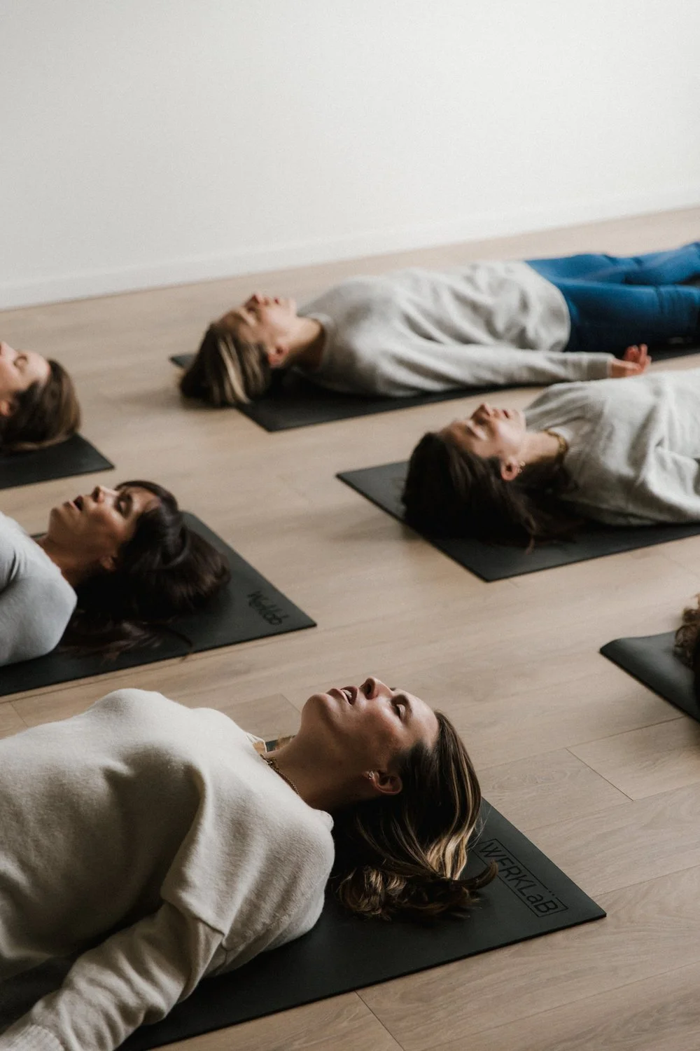 A group of women participating in a yoga class, lying on black mats on a wooden floor.