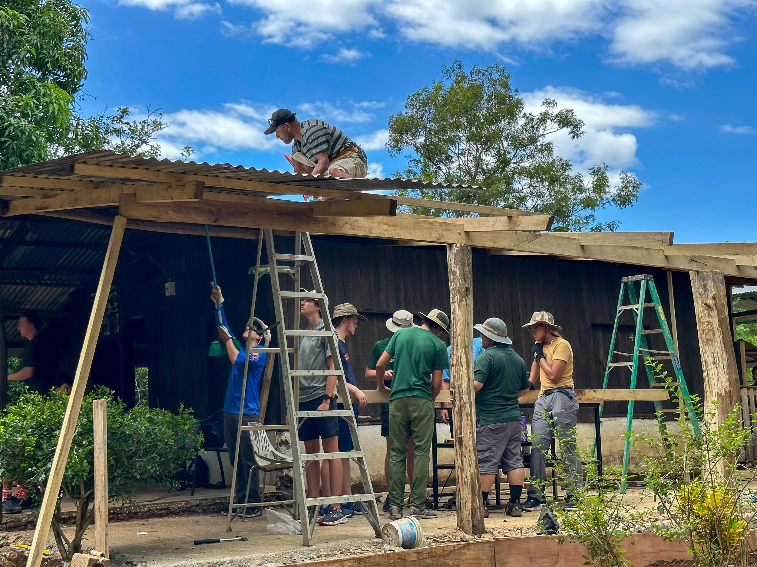 Group of people working on constructing a wooden building outdoors, with some people on ladders and others gathered around, under a blue sky with clouds.